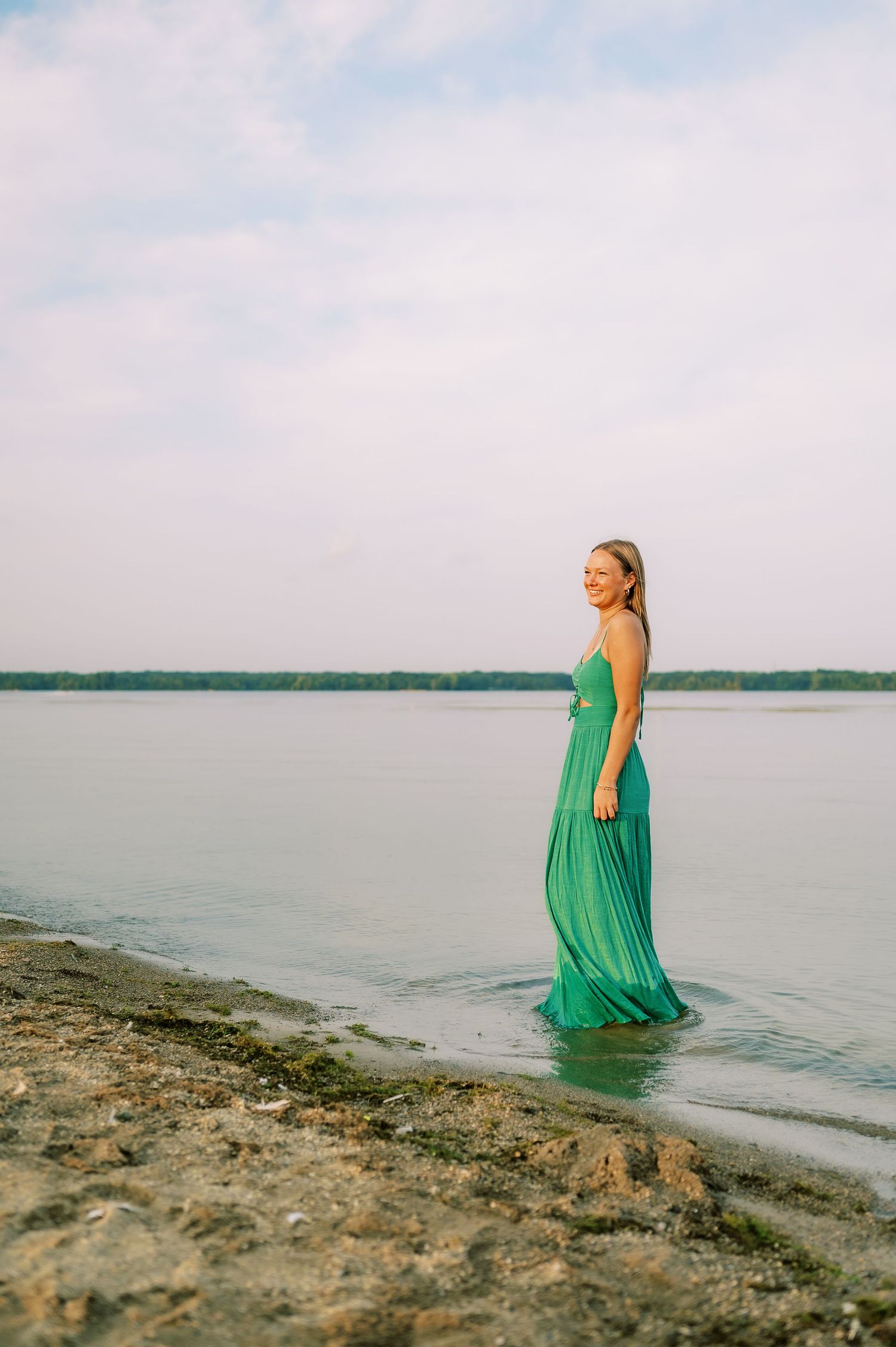 A person in a flowing green dress walks along a rocky shoreline near calm waters at sunset.