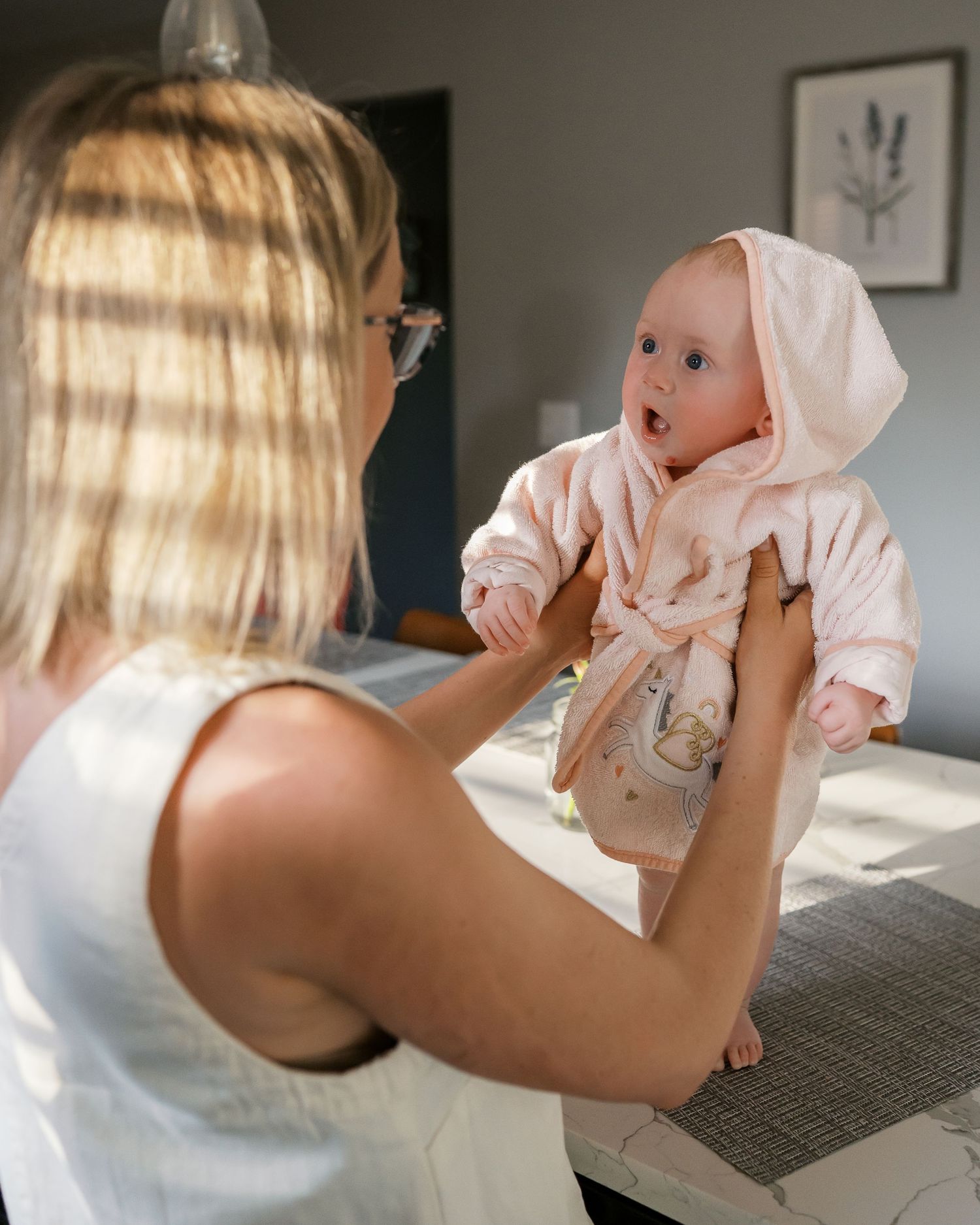 Baby wearing pink hooded outfit being held up in sunlit room.