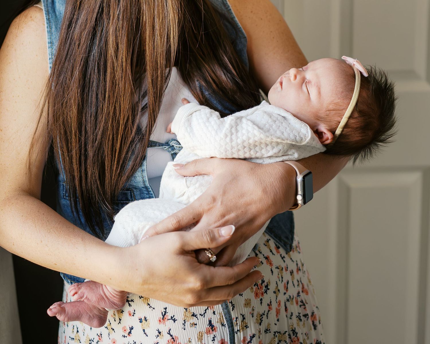 A newborn baby wearing white being cradled in loving arms against a soft interior background.
