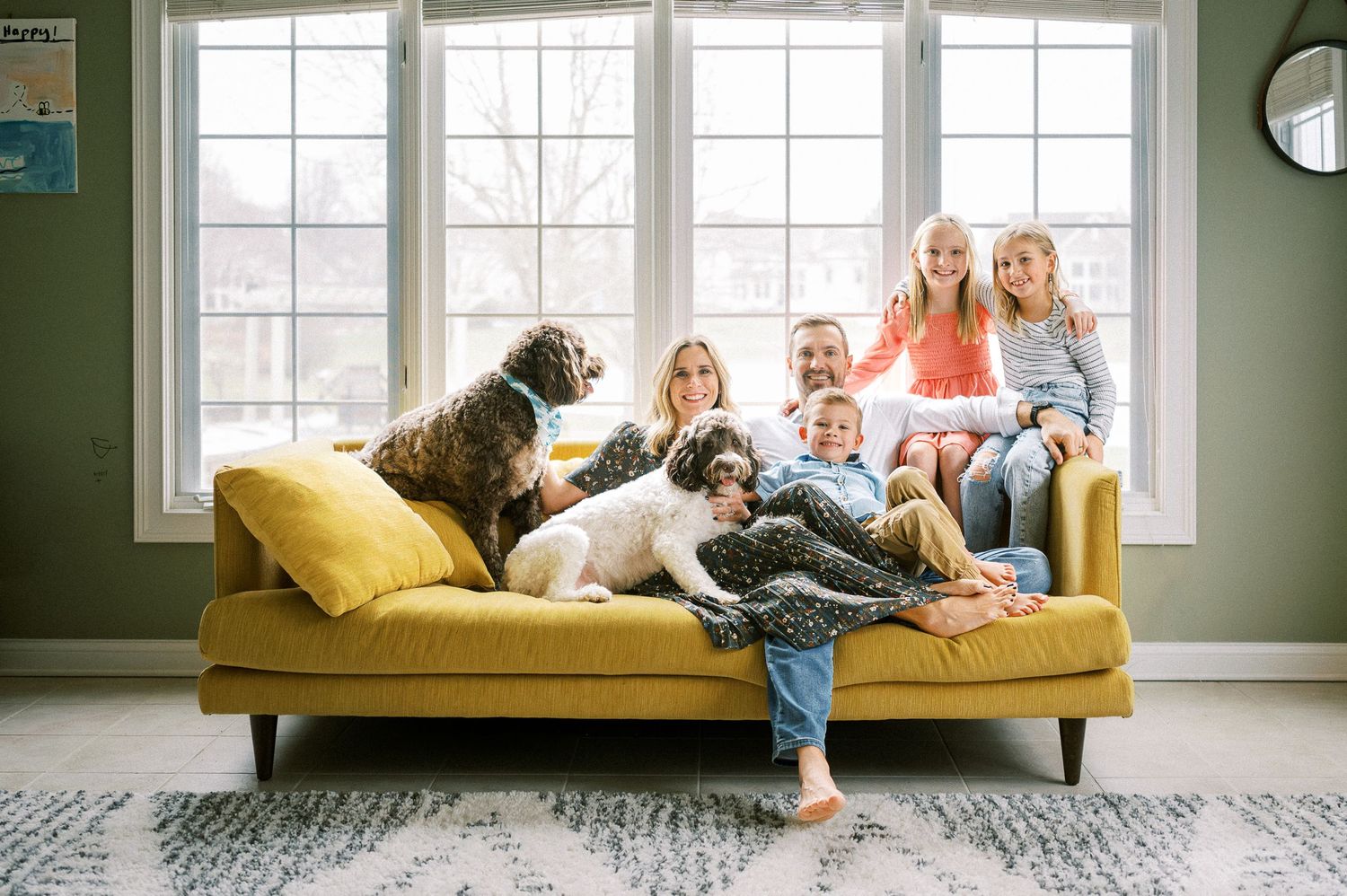 Family relaxing on yellow couch with dogs by bright windows in living room.