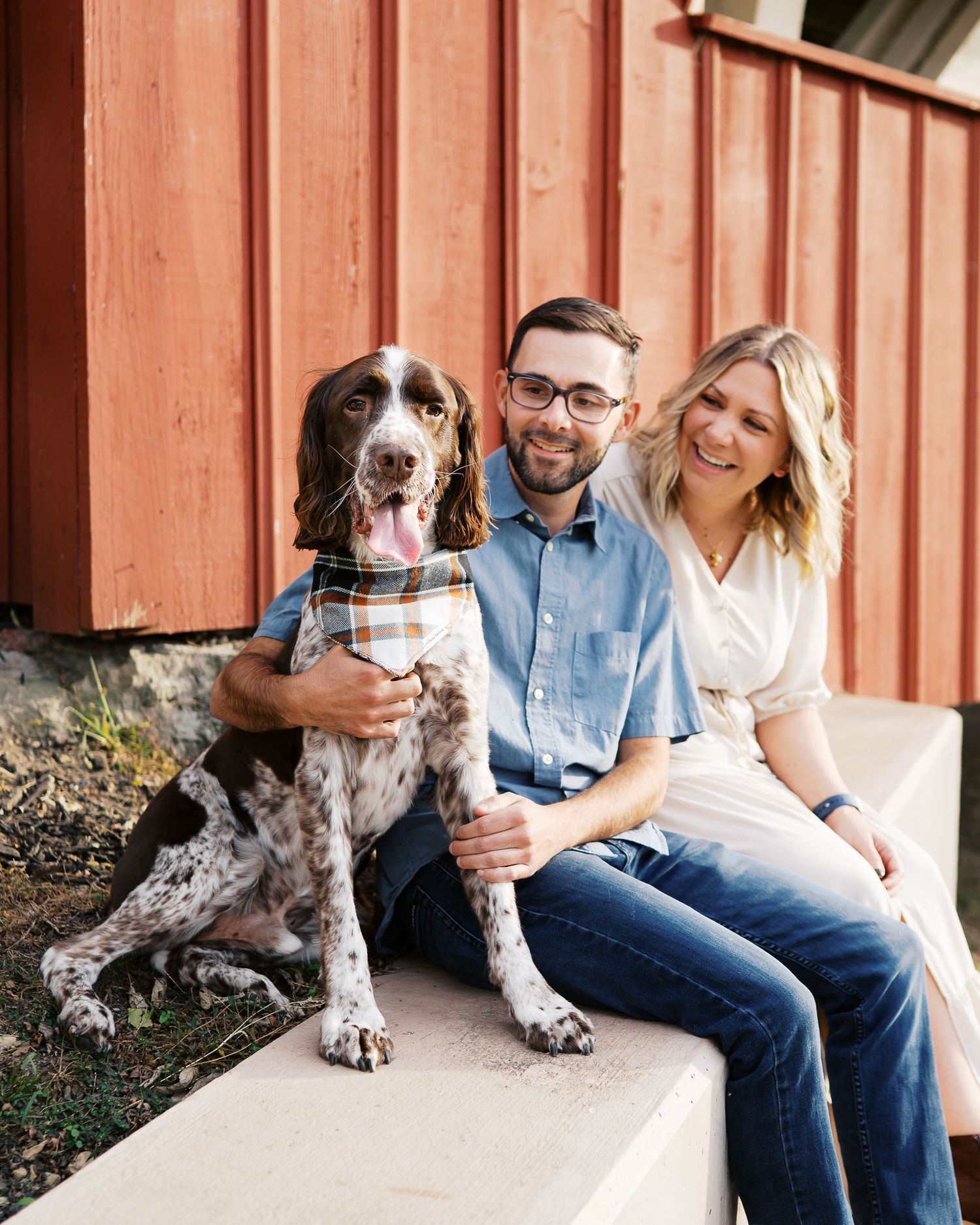 A spotted dog sits with people on a white concrete ledge against a red barn wall.
