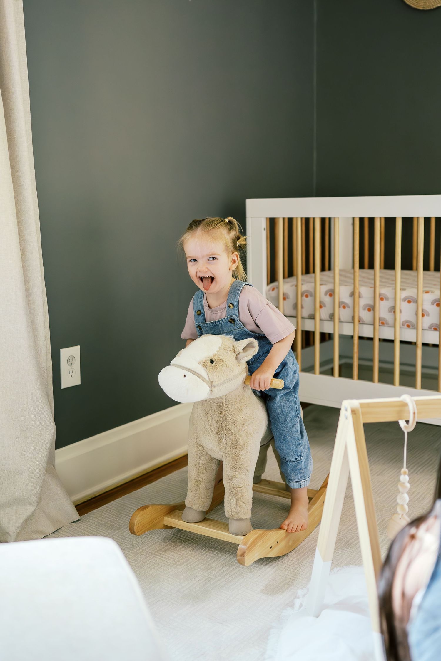 Toddler plays excitedly on plush rocking horse toy near wooden crib against dark green wall.
