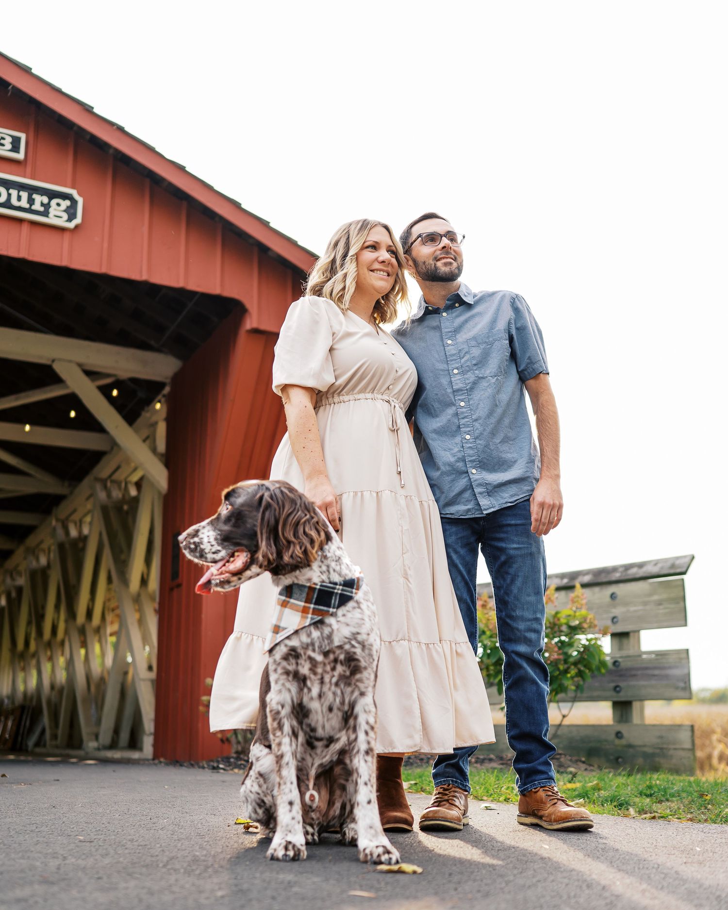 A spotted dog stands near a couple by a covered bridge entrance.