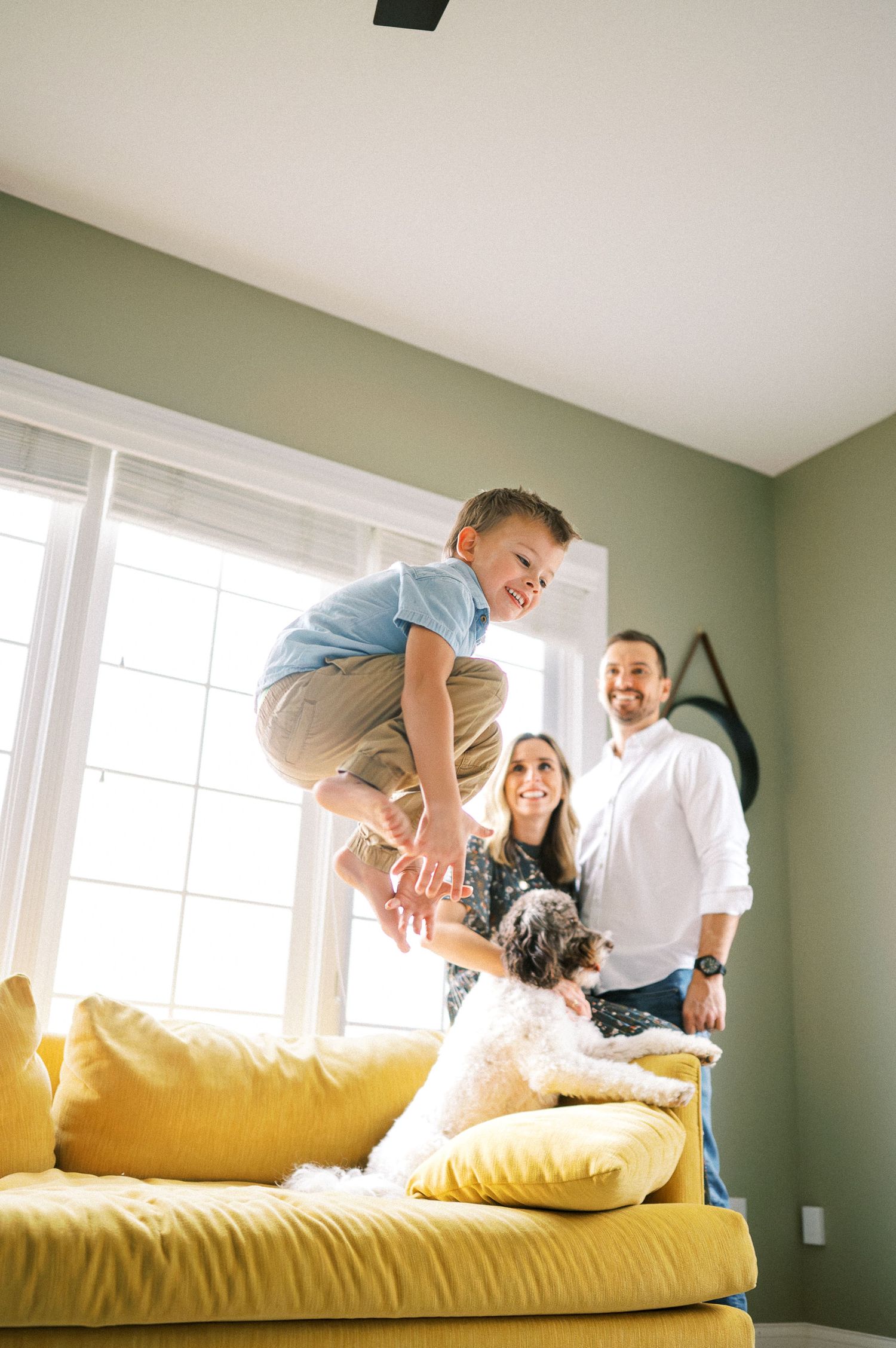 Family members playing and jumping together on a bright yellow couch near large windows.