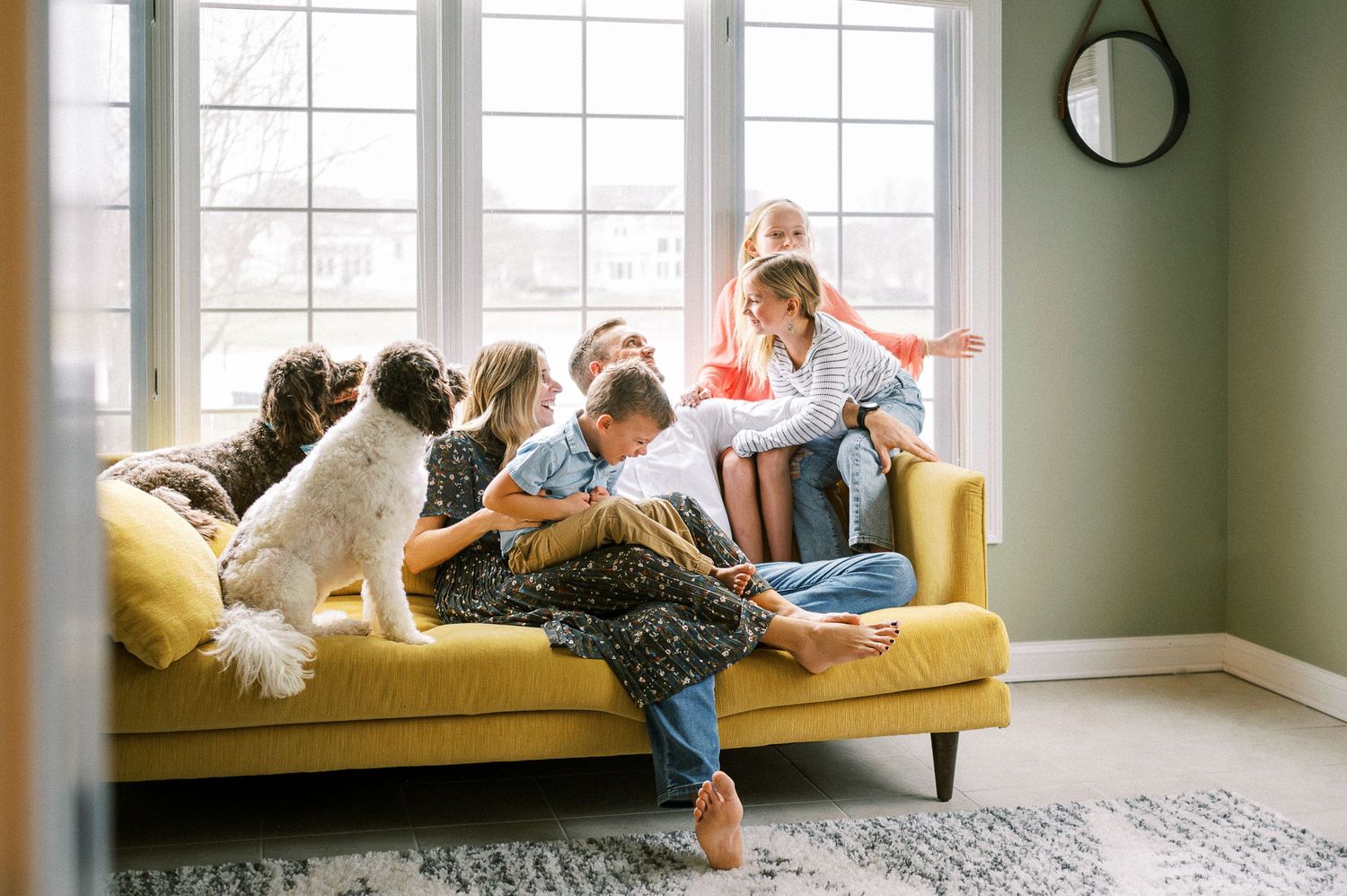 Playful family moment on yellow couch with dogs in sunlit living room.