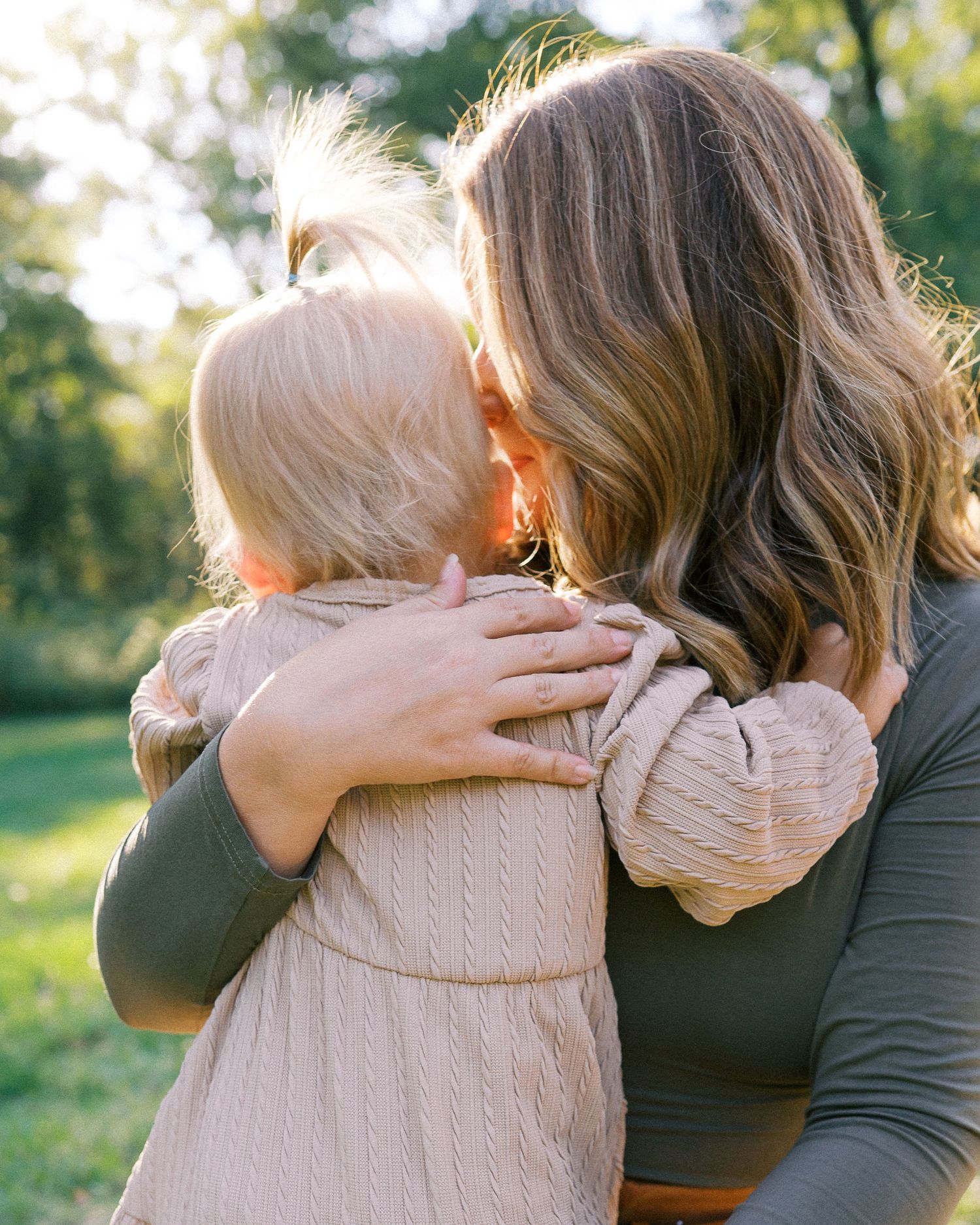 A tender embrace between two people in a sunlit outdoor setting wearing neutral colored clothing.