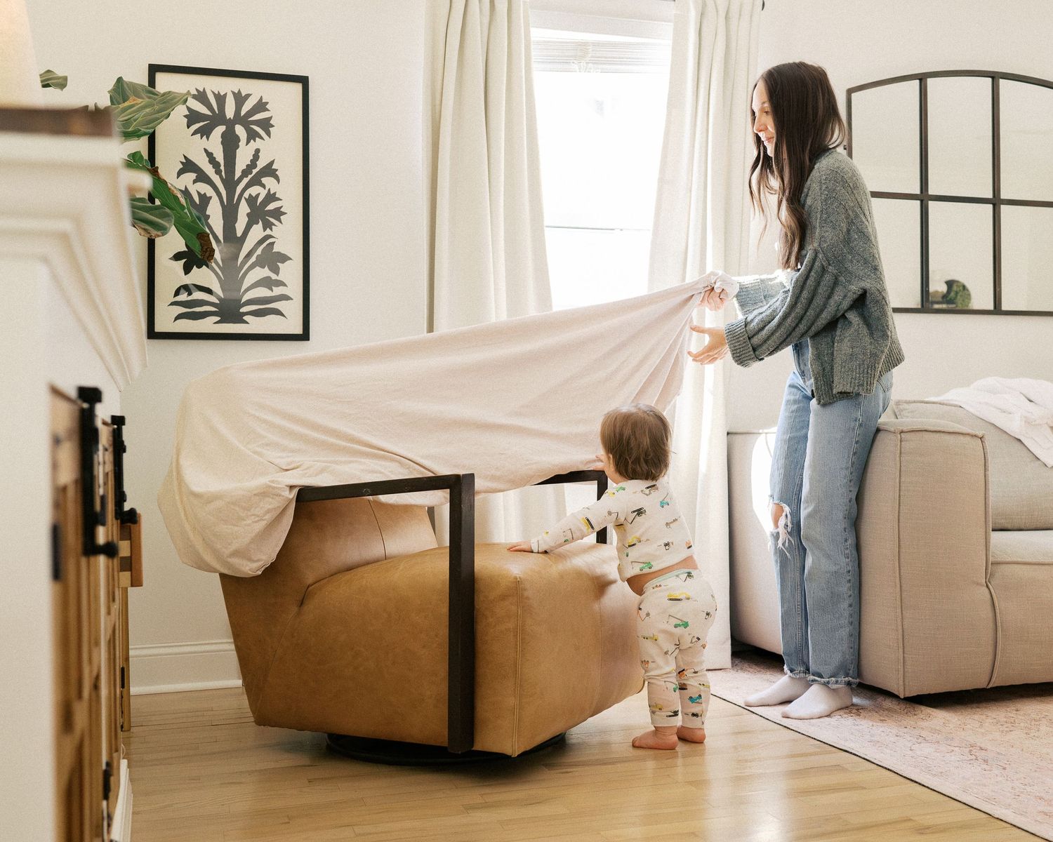 Playing peek-a-boo under a blanket fort in a cozy living room with natural light and botanical artwork.