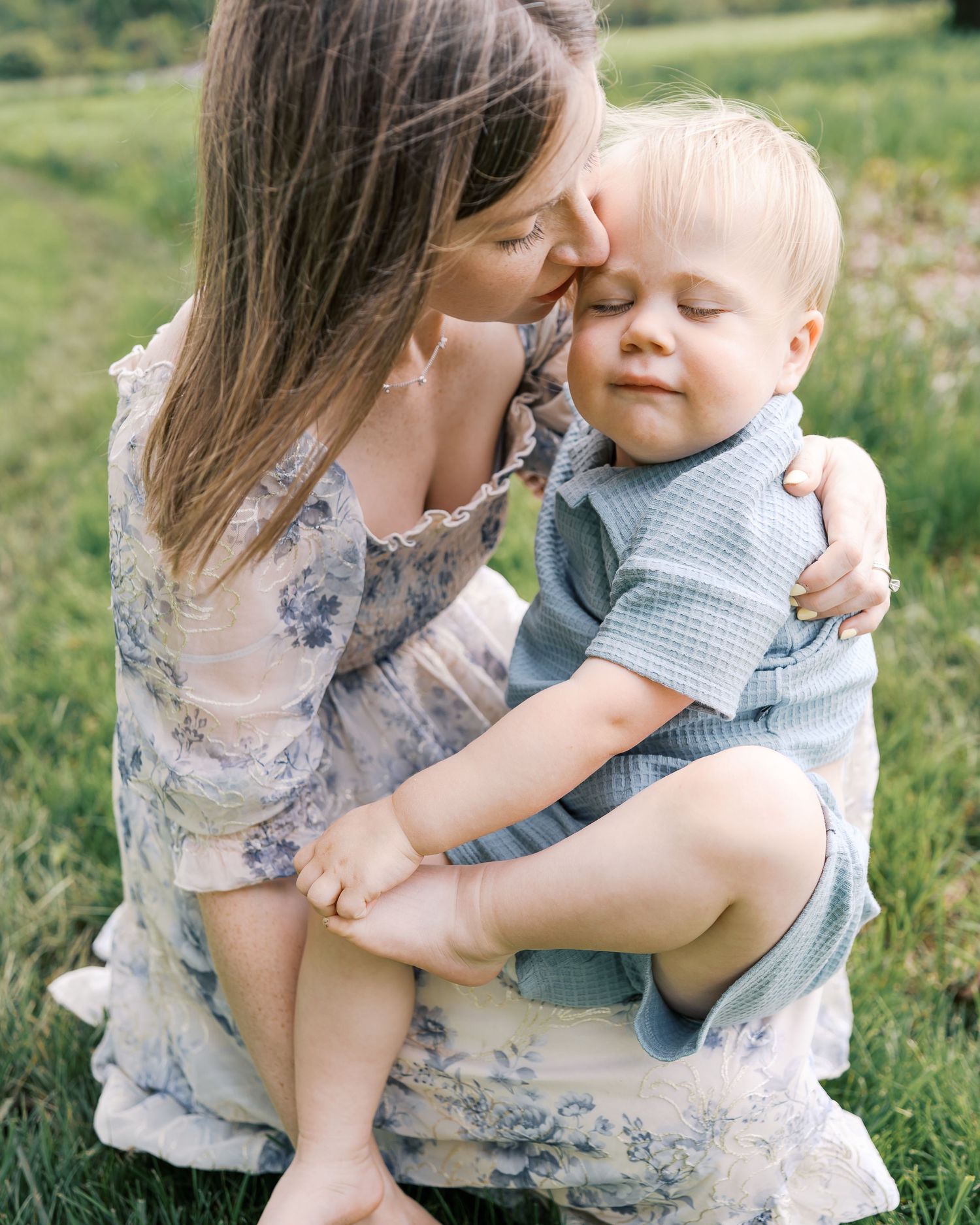 Tender moment between parent and child sitting in a green meadow wearing floral and checkered clothing.