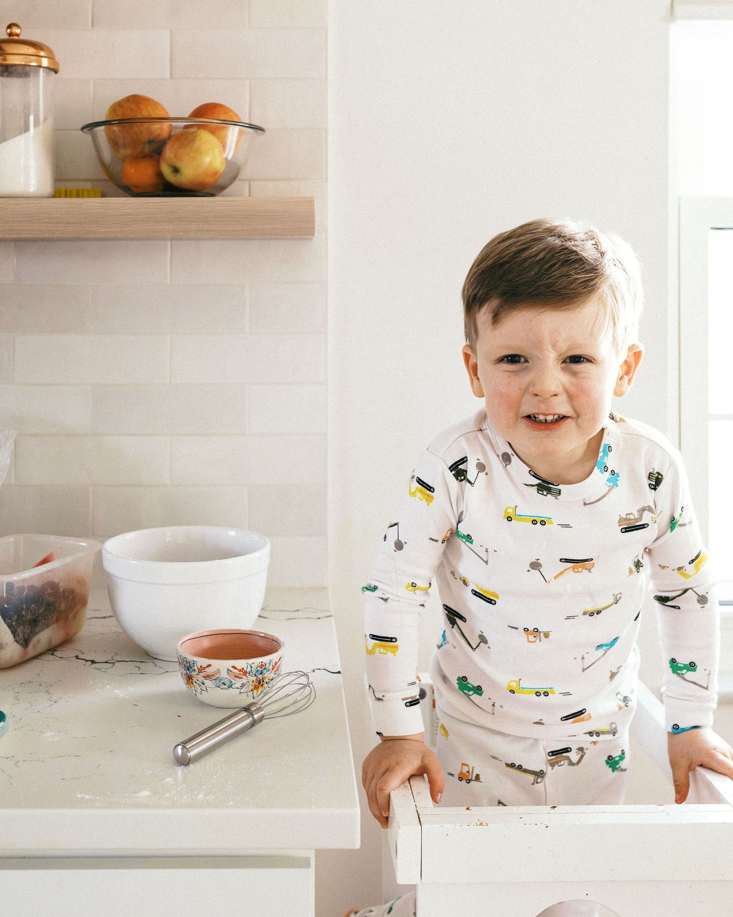 A young child in vehicle-patterned pajamas smiling while standing at a kitchen counter.