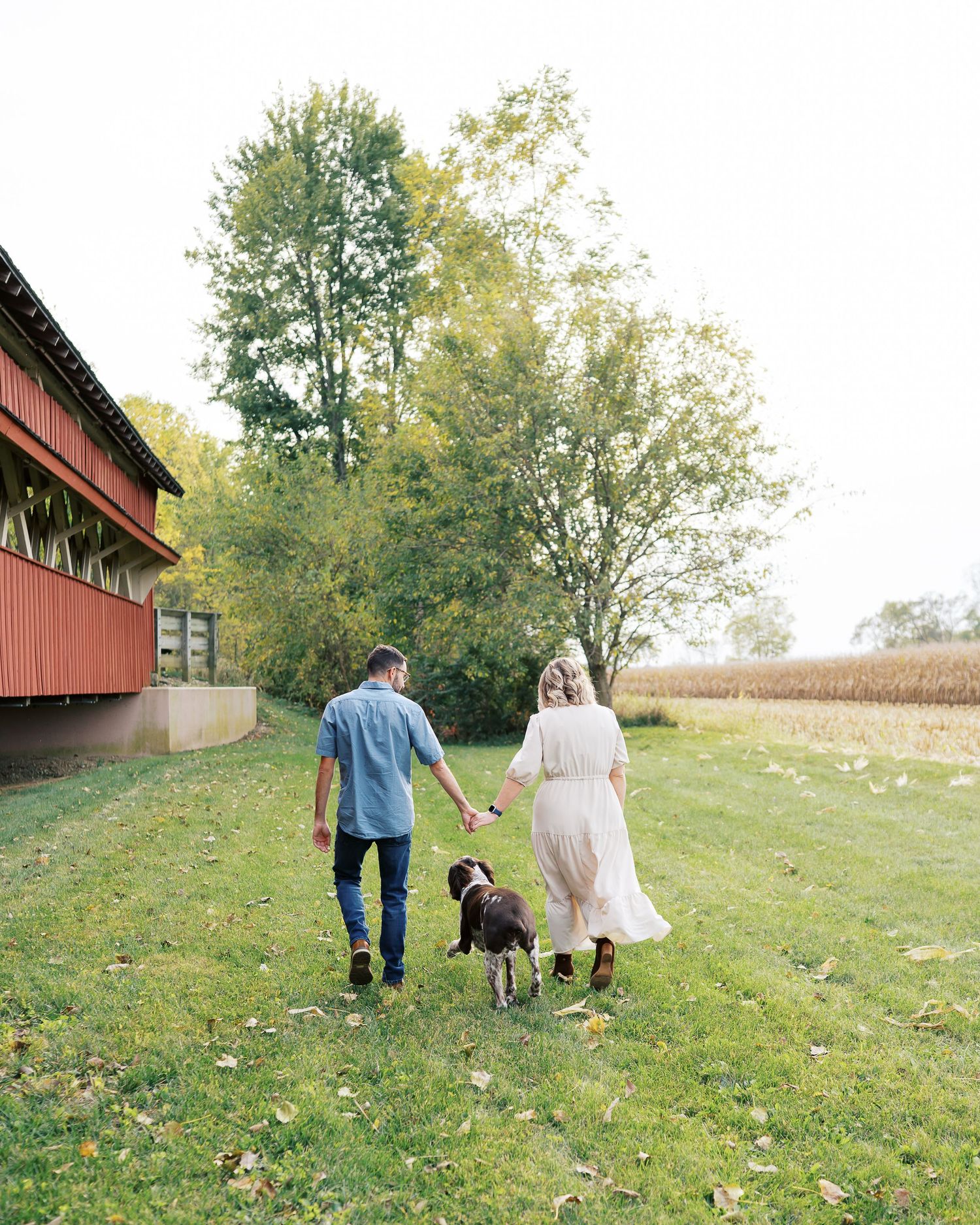 A couple and their dog walk together on a grassy hillside near a red barn on an autumn day.