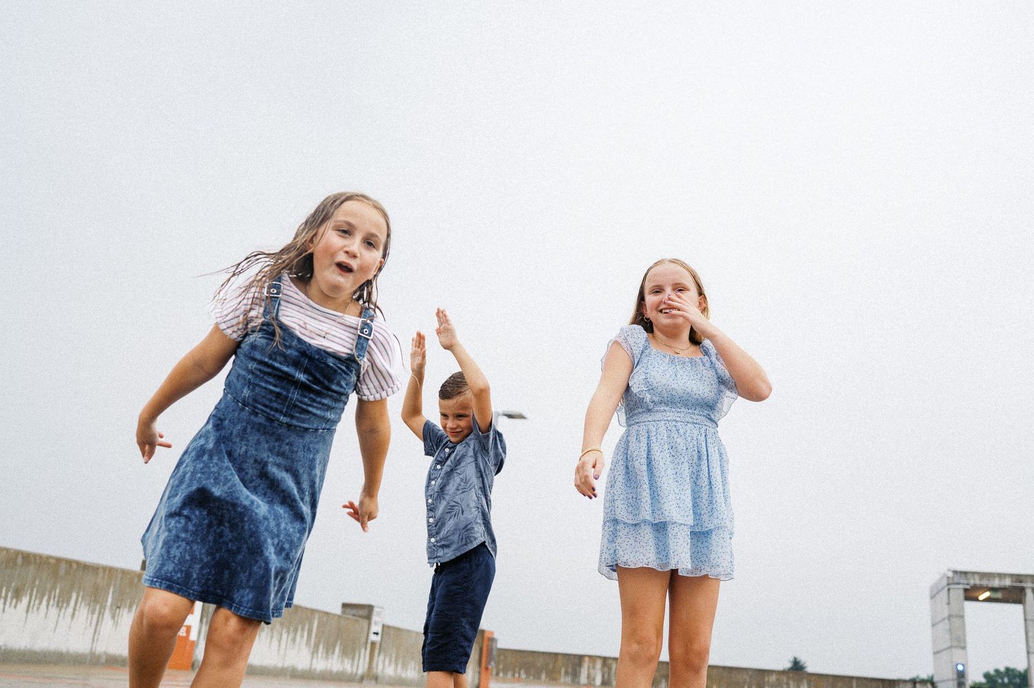 Three children play and jump together on a boardwalk by the water on an overcast day.