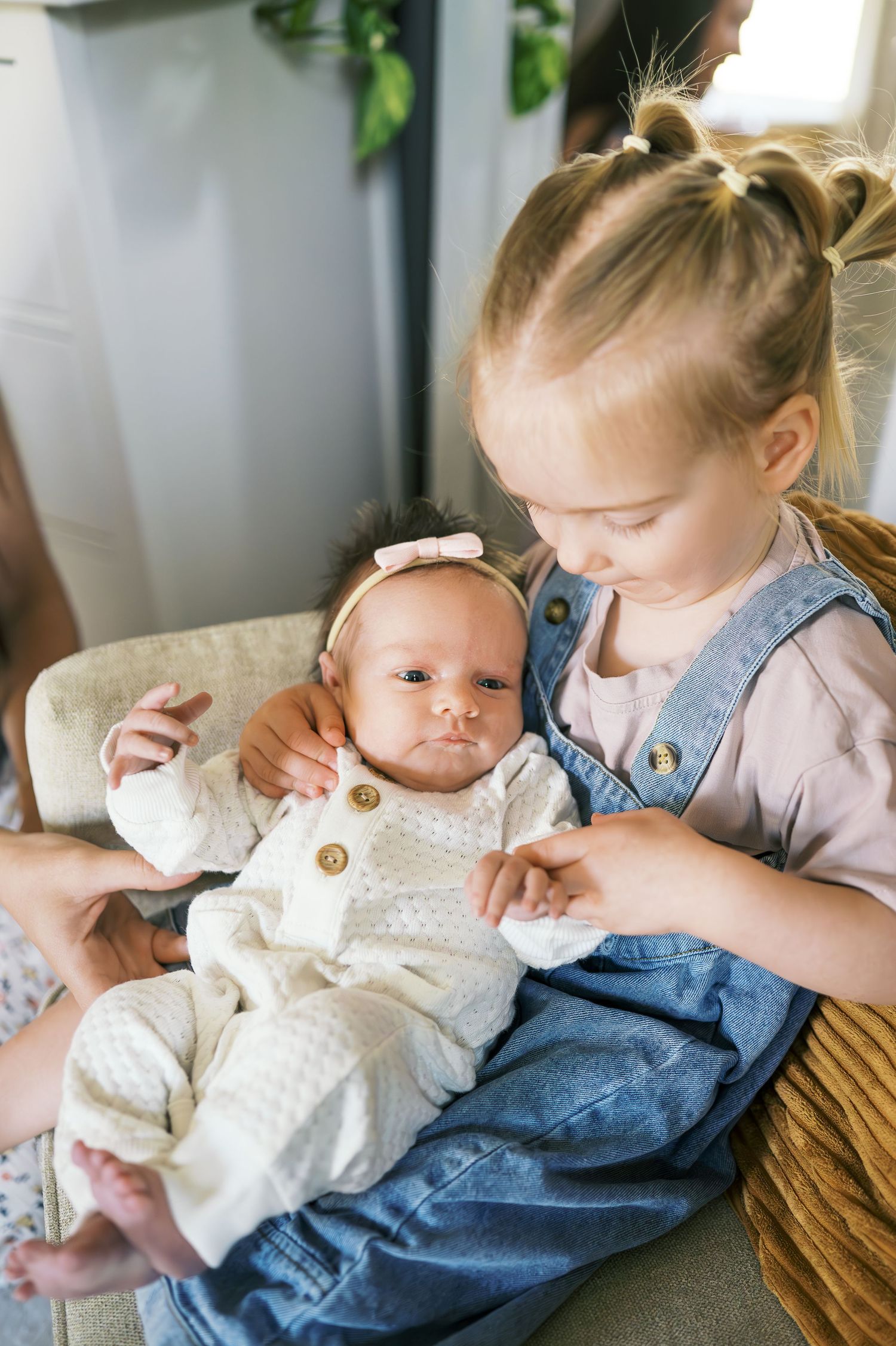 Two children sit together on brown chair with one wearing denim overalls.