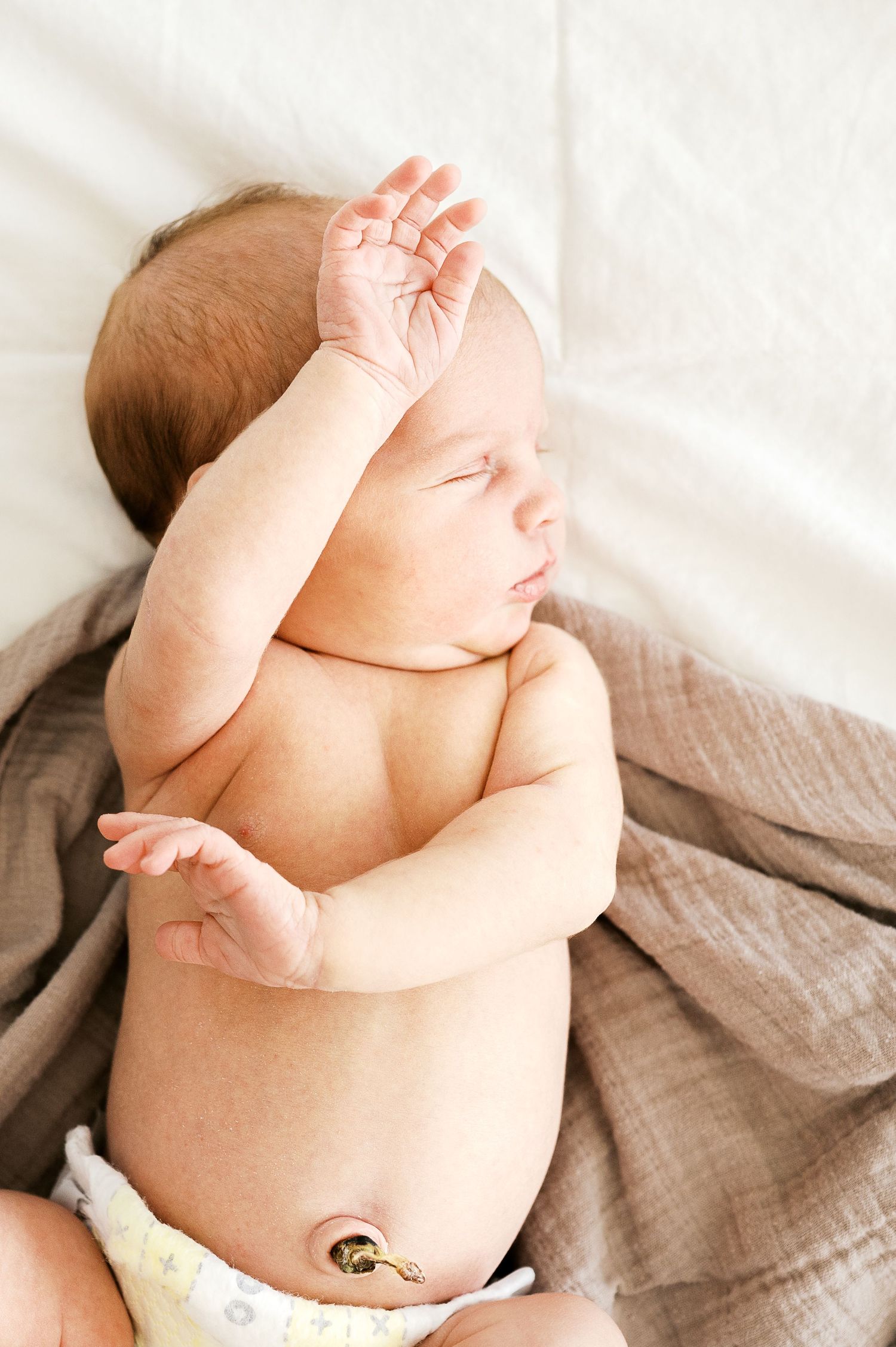 A sleeping newborn baby lies on soft fabric with one hand raised to their head.