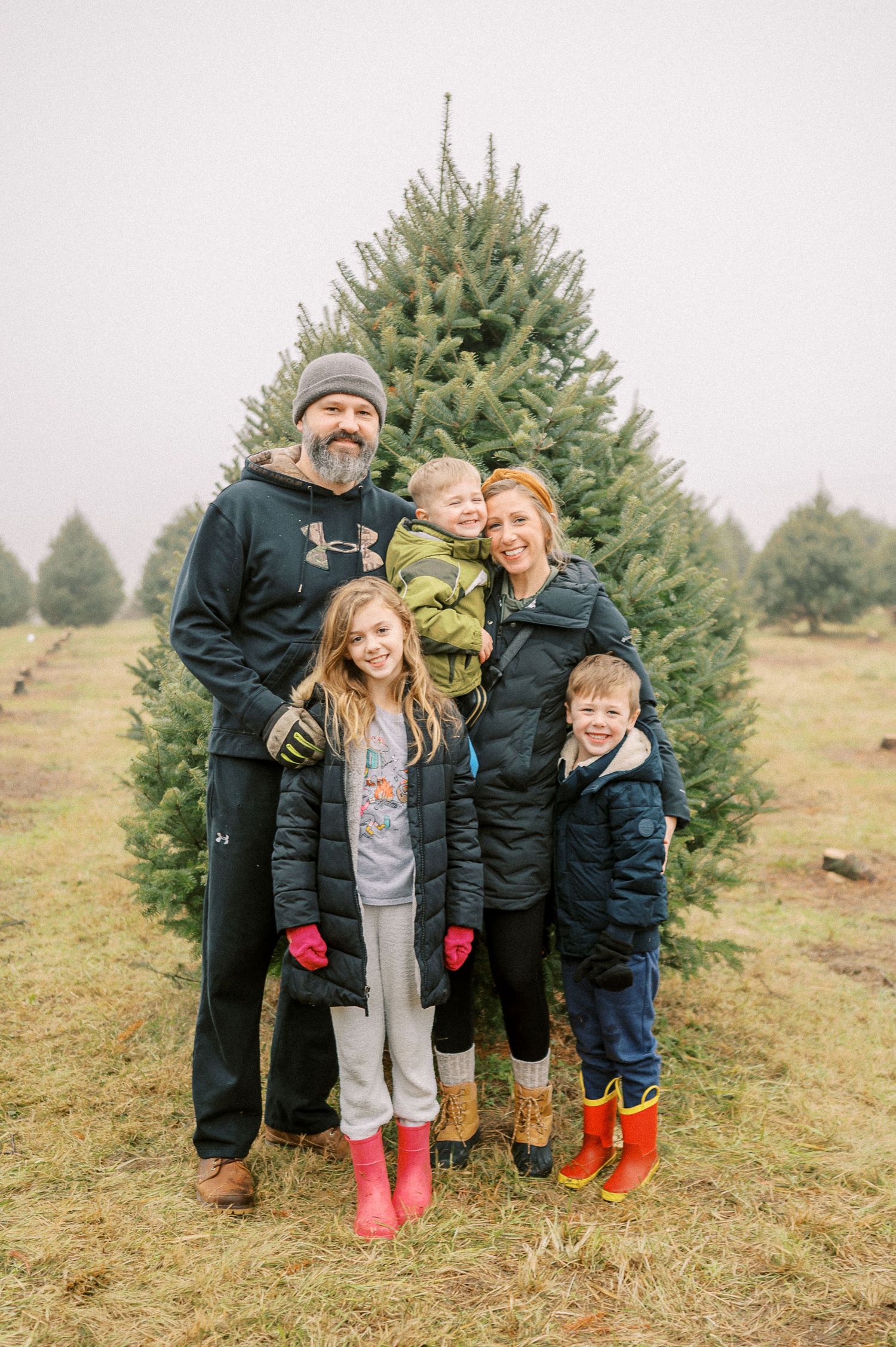 Family wearing warm winter clothing poses together at a Christmas tree farm on an overcast day.