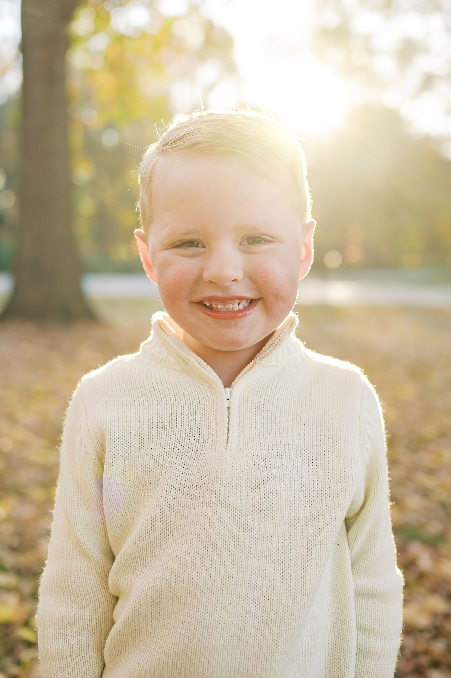 Smiling child wearing cream knit sweater standing outdoors in autumn sunlight.