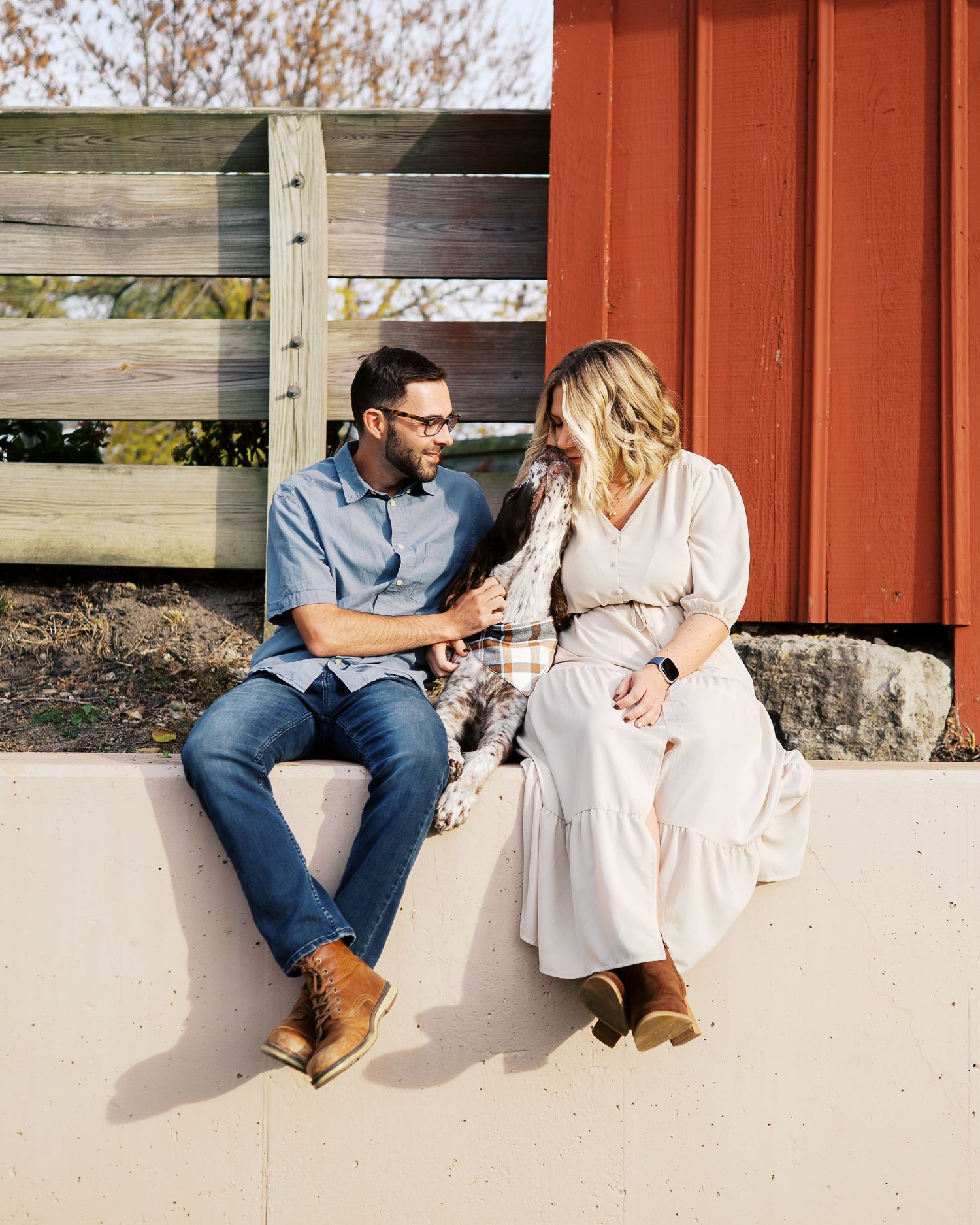 A couple shares a tender moment while sitting together against a red barn wall on a sunny day.