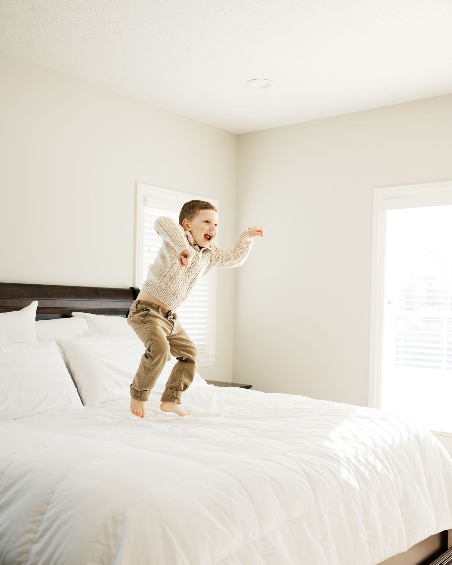 Child jumping with excitement on white bed in bright bedroom.
