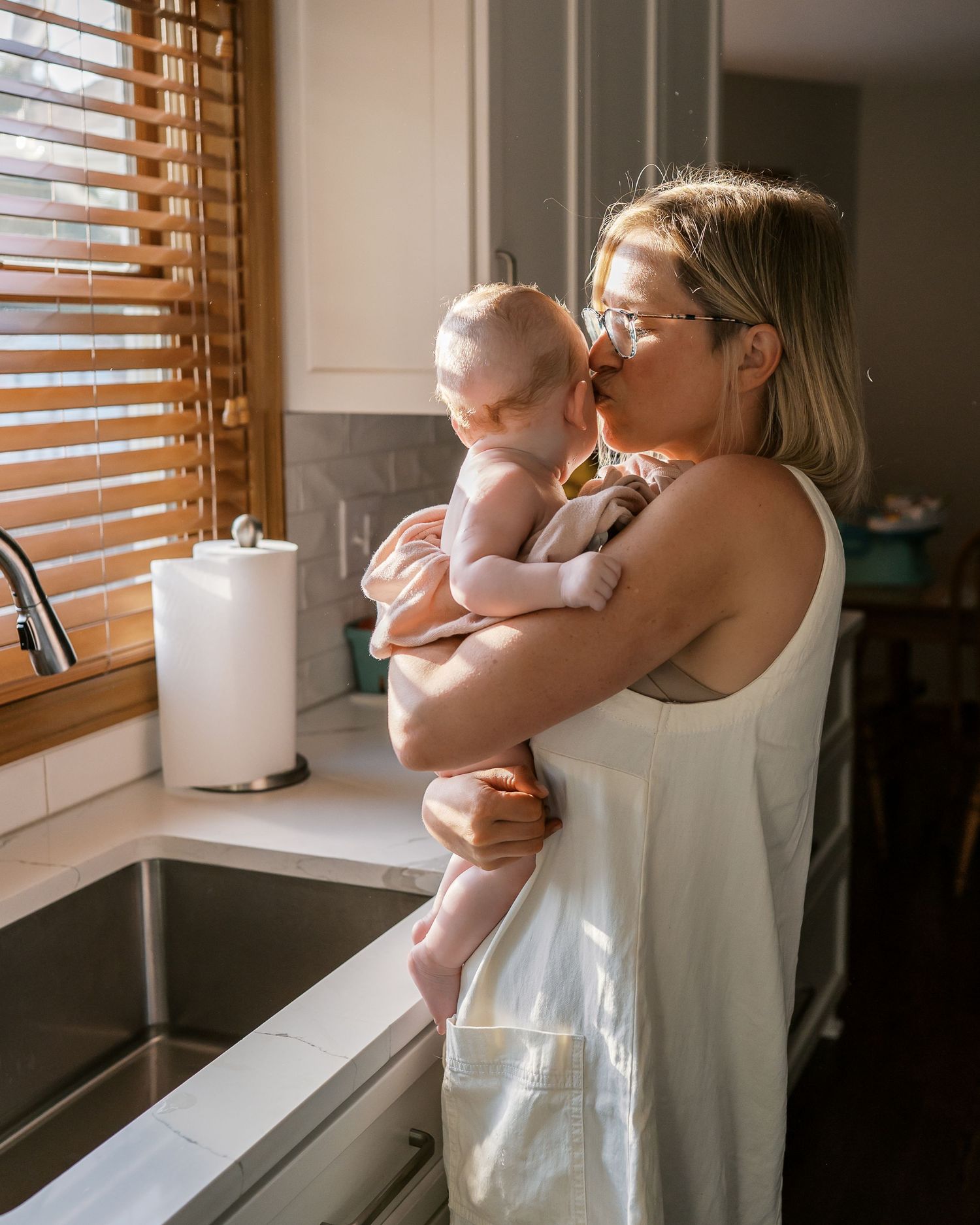 Intimate kitchen scene with a baby being held near a window with wooden blinds.