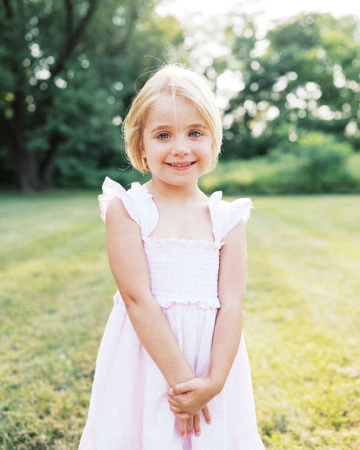 A child wearing a white dress poses playfully in a sunny outdoor garden setting.
