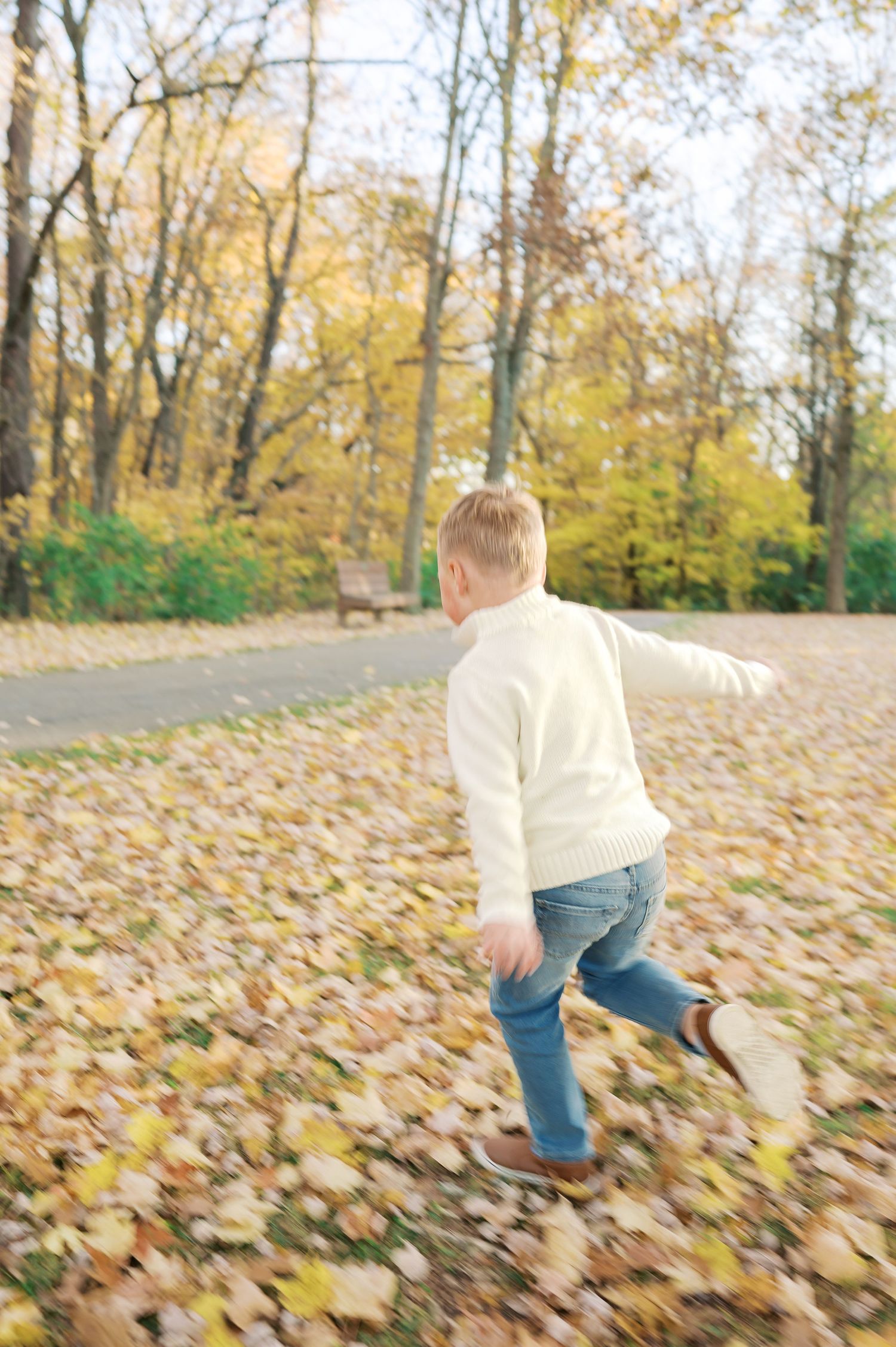 Child in white sweater and jeans running through fallen leaves in autumn park.