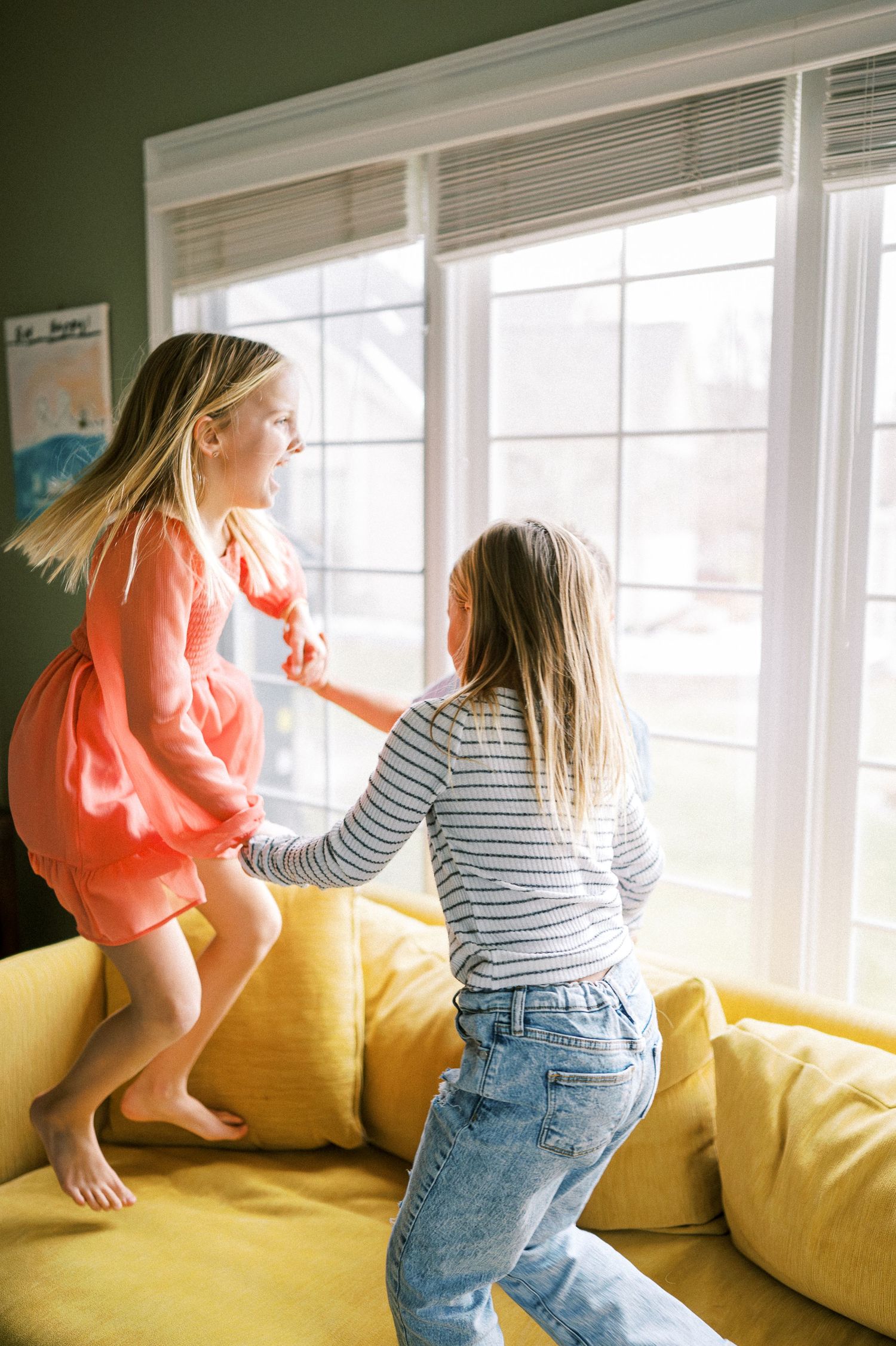 Two people playing and jumping energetically on a yellow couch by large windows.