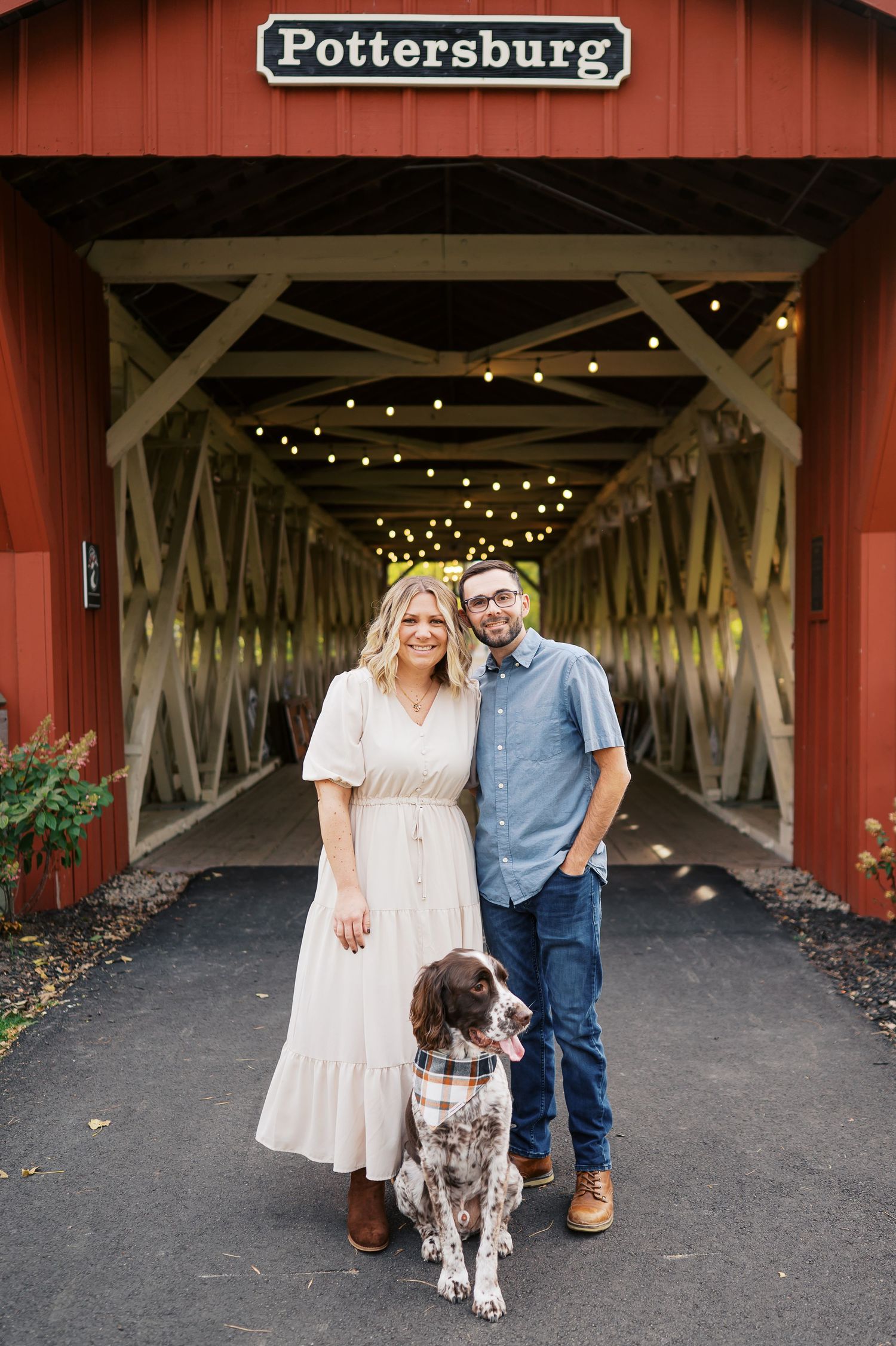 A couple and their spotted dog pose in front of a red covered bridge entrance with string lights in Pottersburg.