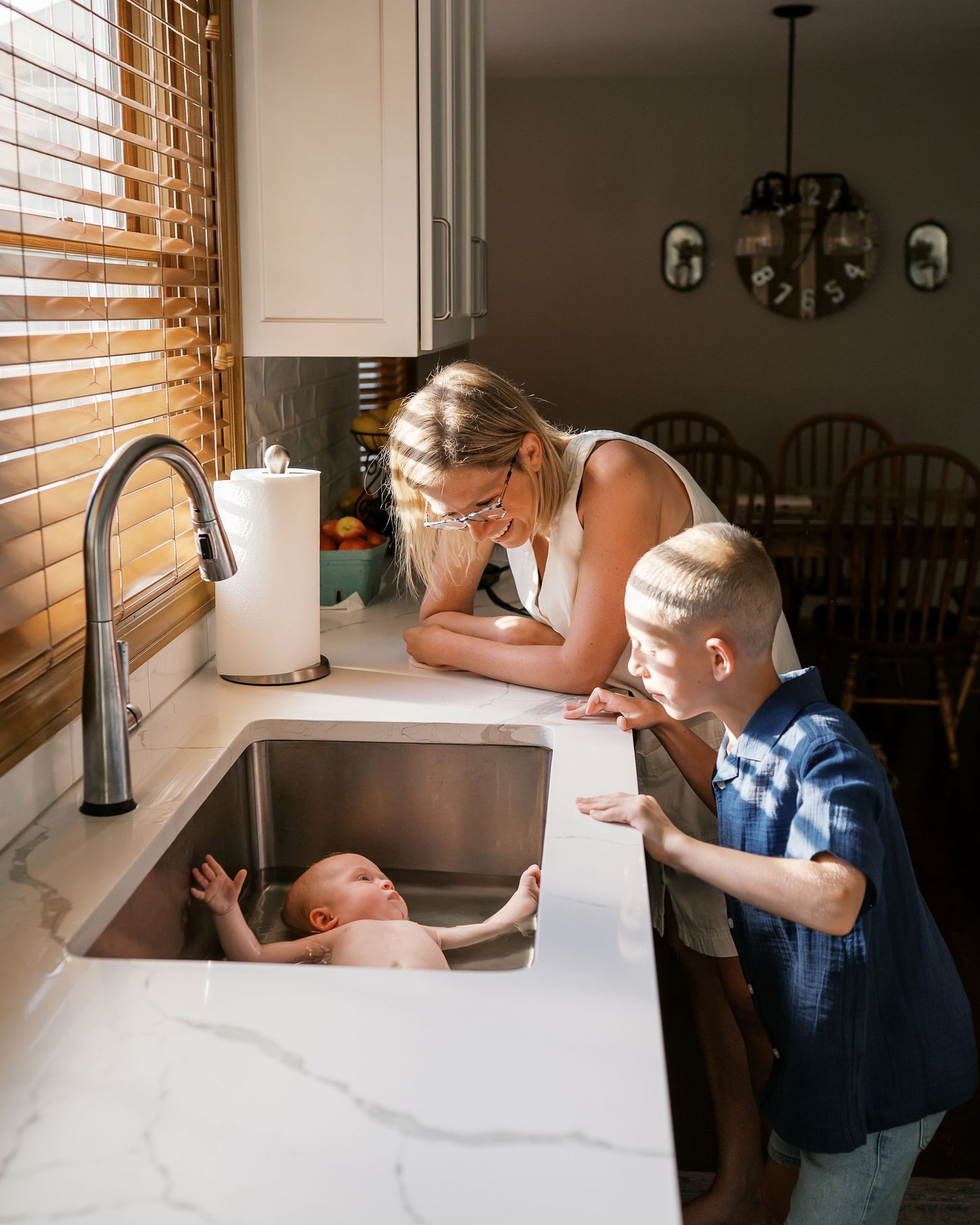 Two people help give baby bath in white marble kitchen counter sink with natural light.