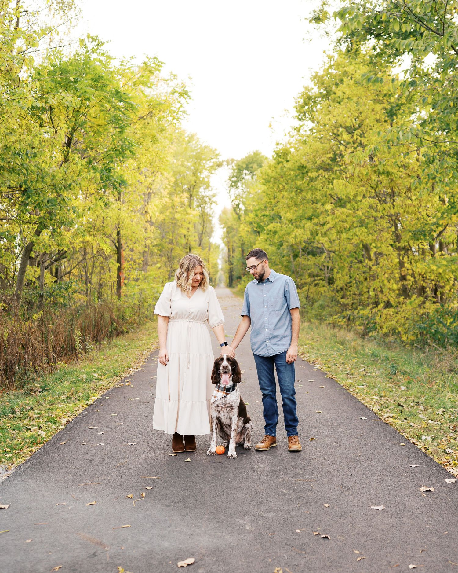 A couple and their spotted dog pose together on a tree-lined path during fall.