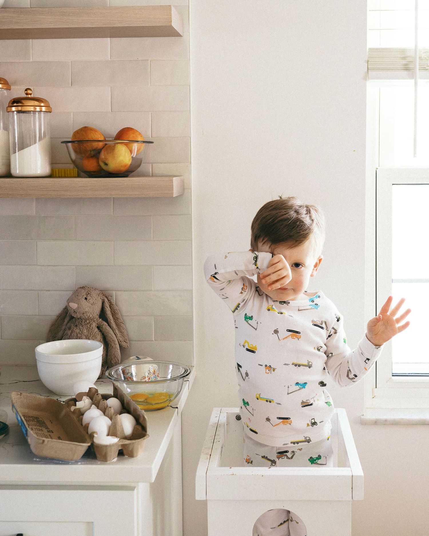 A child in patterned pajamas standing on a learning tower next to kitchen shelves with fruit.