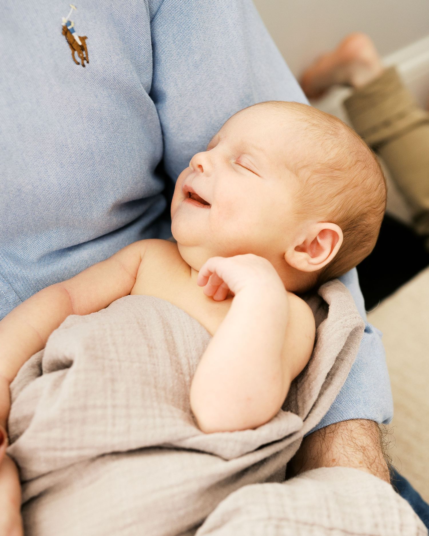 A sleeping newborn baby rests against a light blue polo shirt with their tiny arm raised.