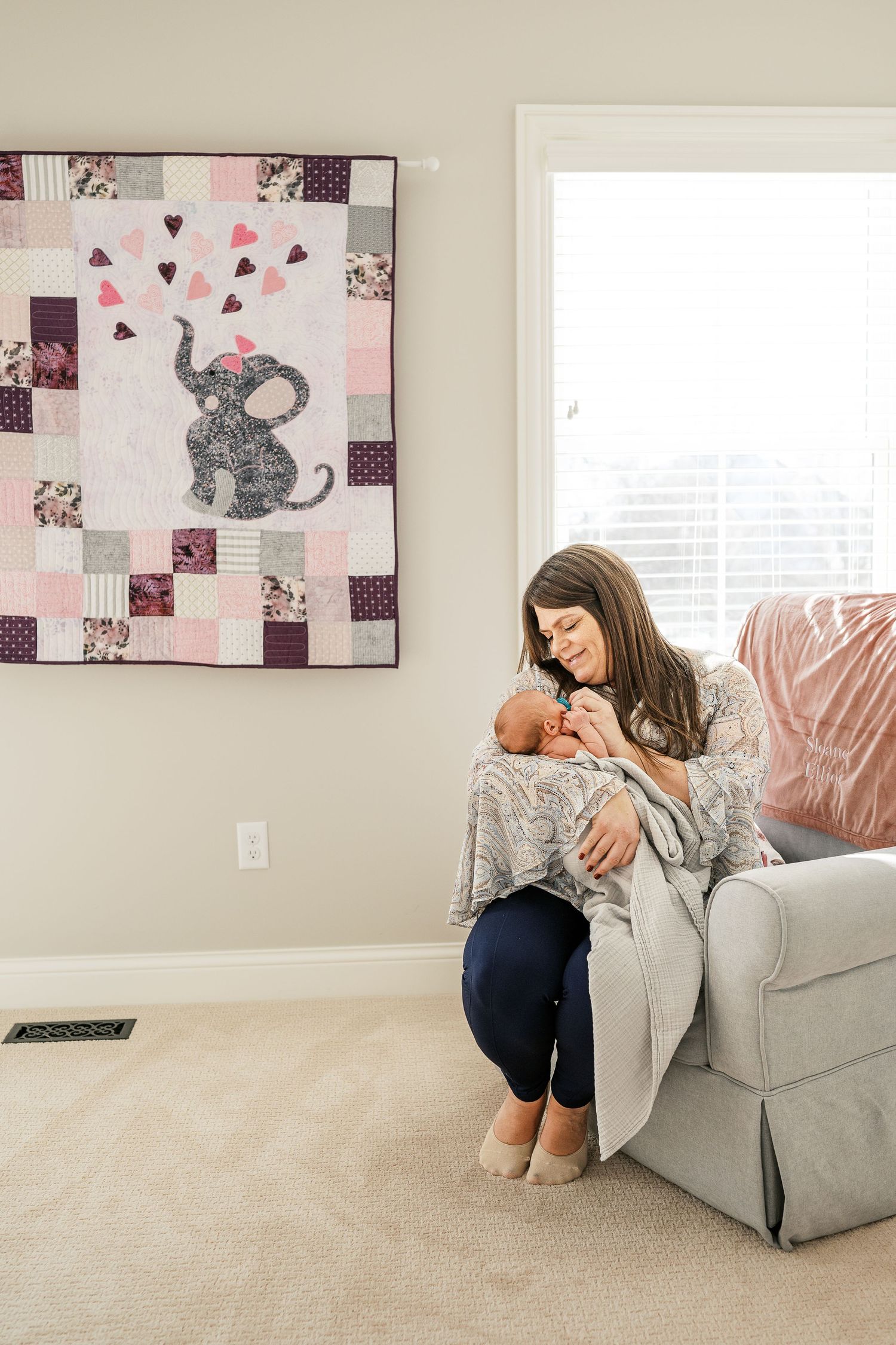 Nursery scene with elephant artwork and parent holding baby in gray armchair.