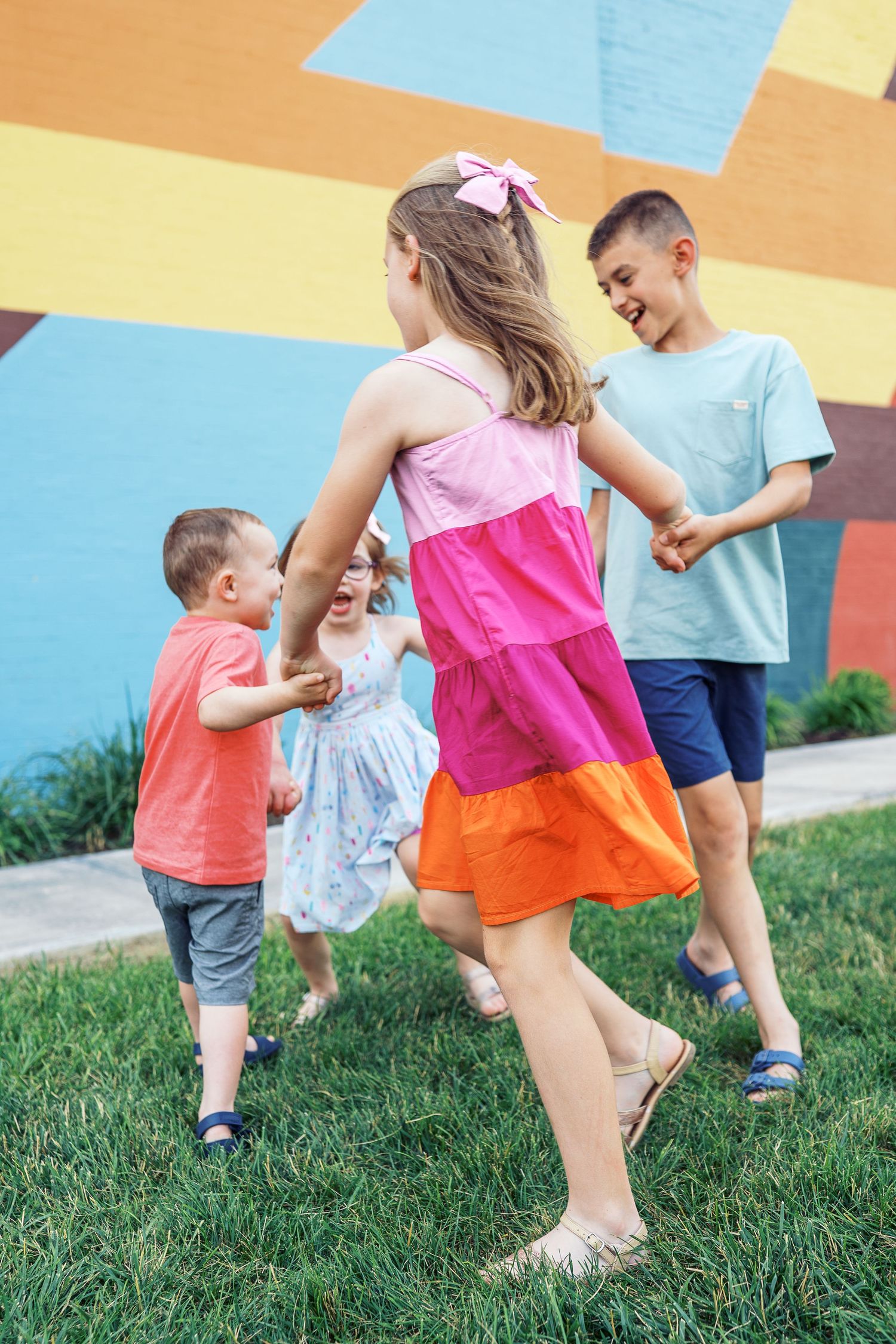 Running children play together on green grass against a colorful wall backdrop during an outdoor activity.