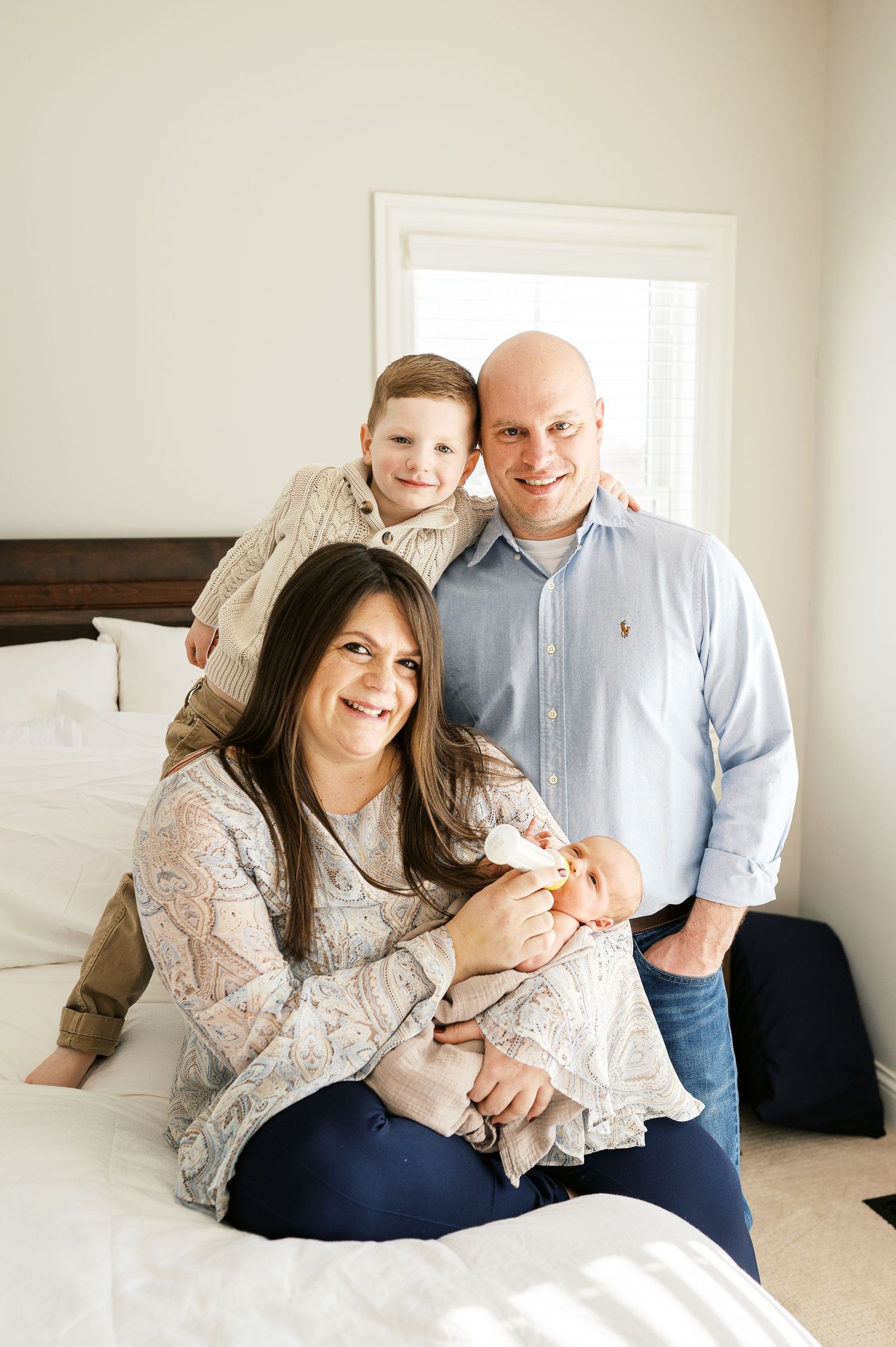 A family gathers on a white bed for an intimate portrait in a bright bedroom.