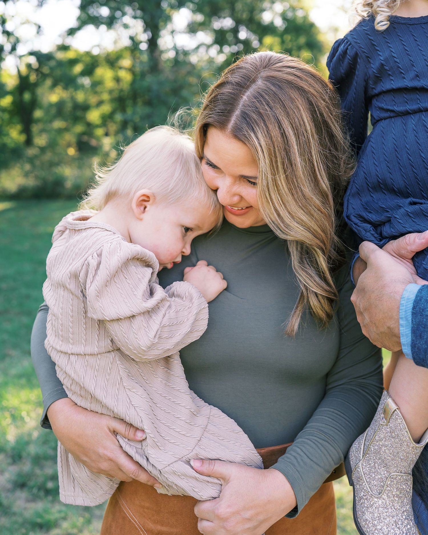 A tender moment between parent and baby in beige sweater against a sunlit outdoor backdrop.