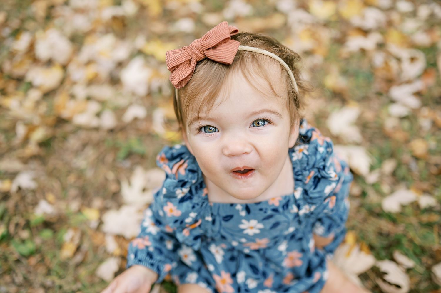 Baby wearing floral dress and bow smiles while sitting among fallen autumn leaves.