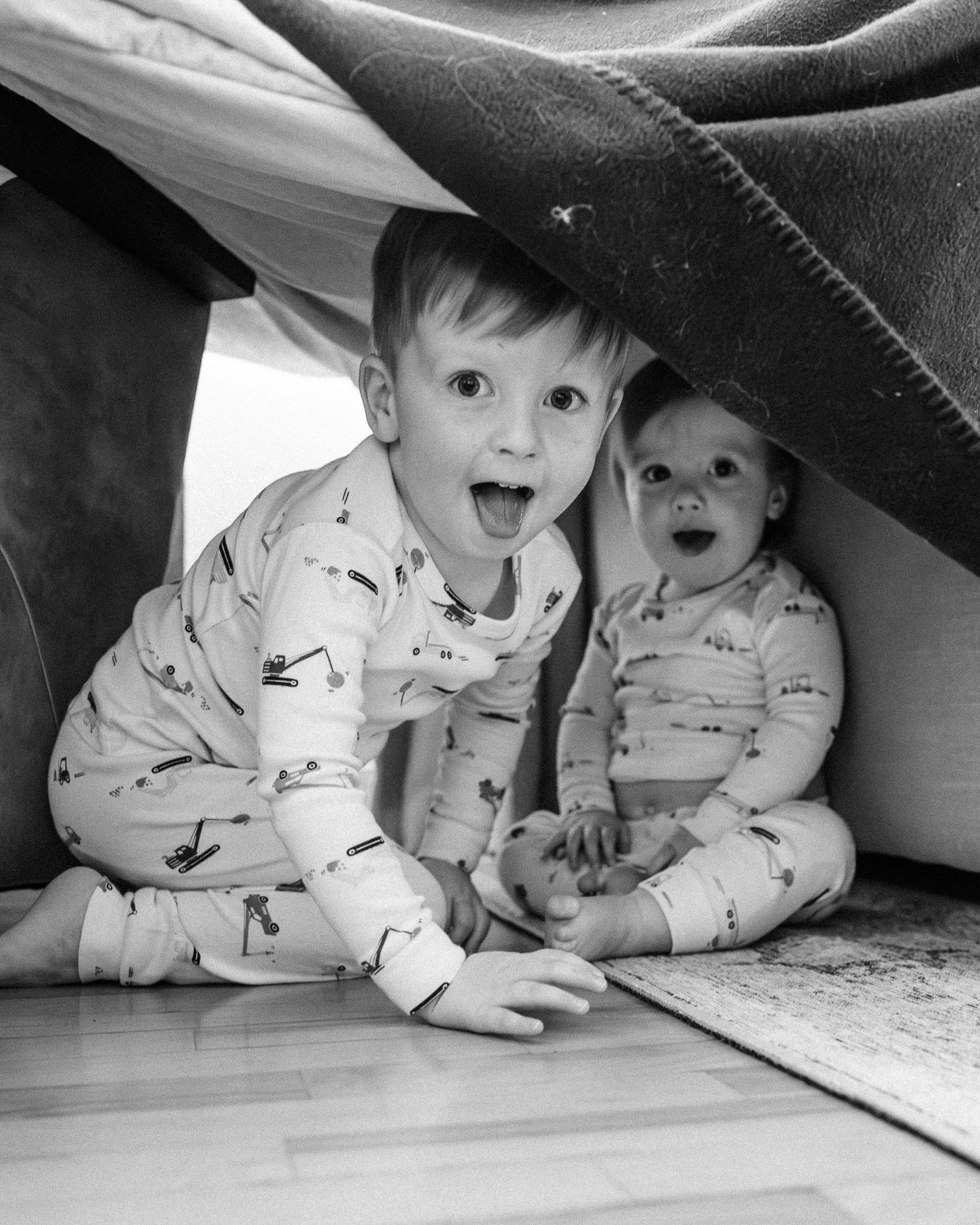 Two small children in matching pajamas playing under a blanket fort on hardwood floor.