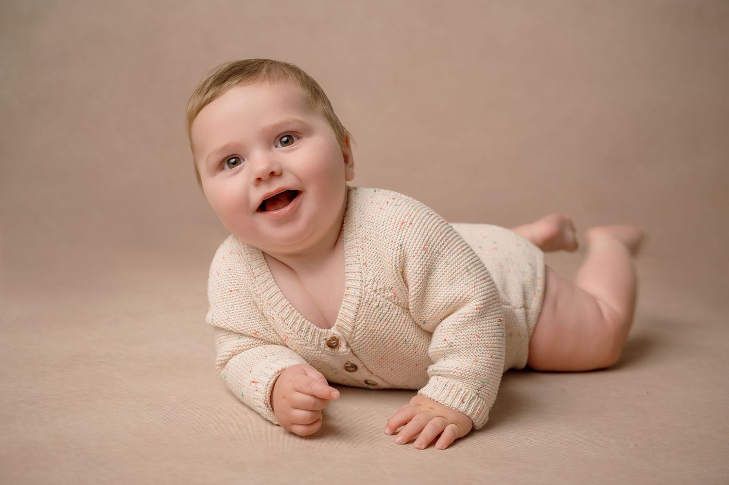 Sweet baby in cream sweater lying on beige backdrop with bright smile.