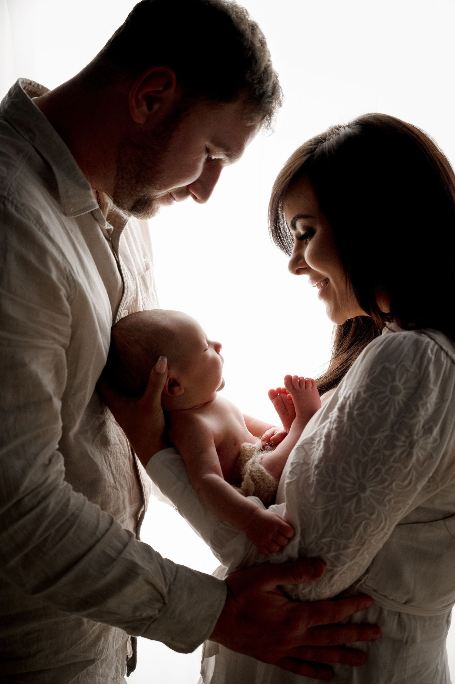 Intimate newborn photography with parents holding their baby in a bright silhouette setting.