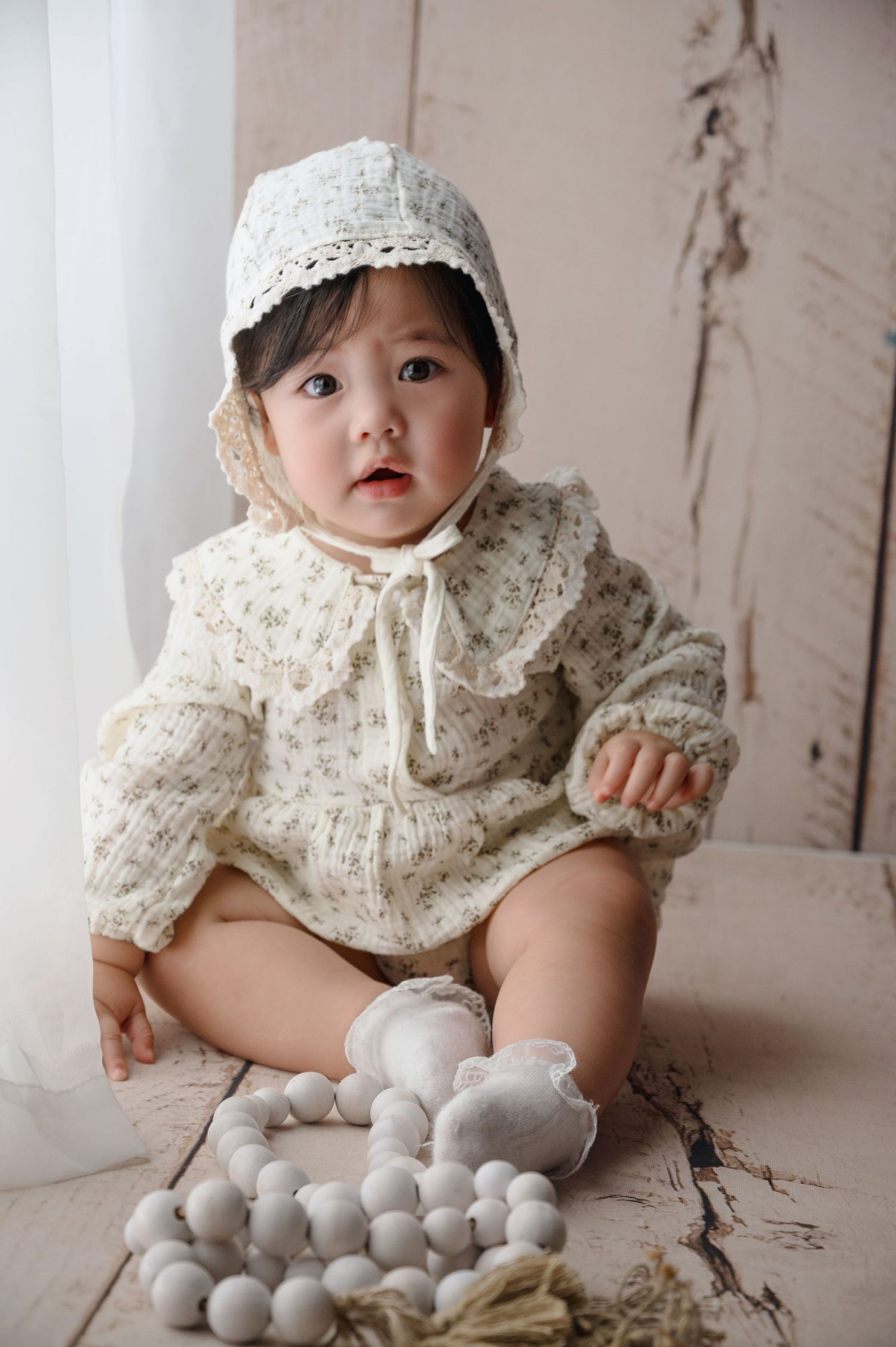 Baby in vintage-style white lace outfit and bonnet sitting on wooden floor with decorative accessories.