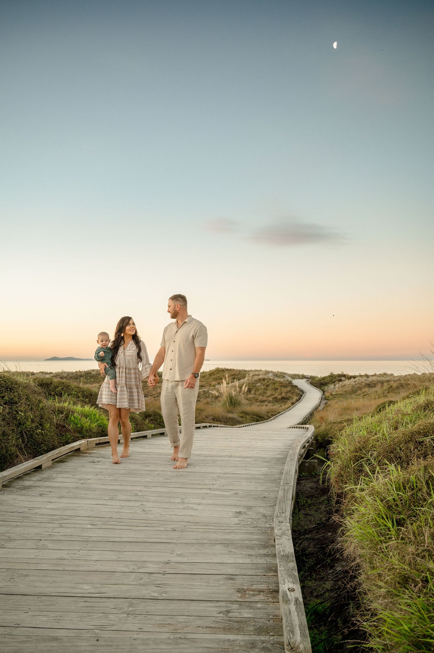 A couple walks along a wooden boardwalk path through coastal dunes at sunset with a crescent moon in the sky.