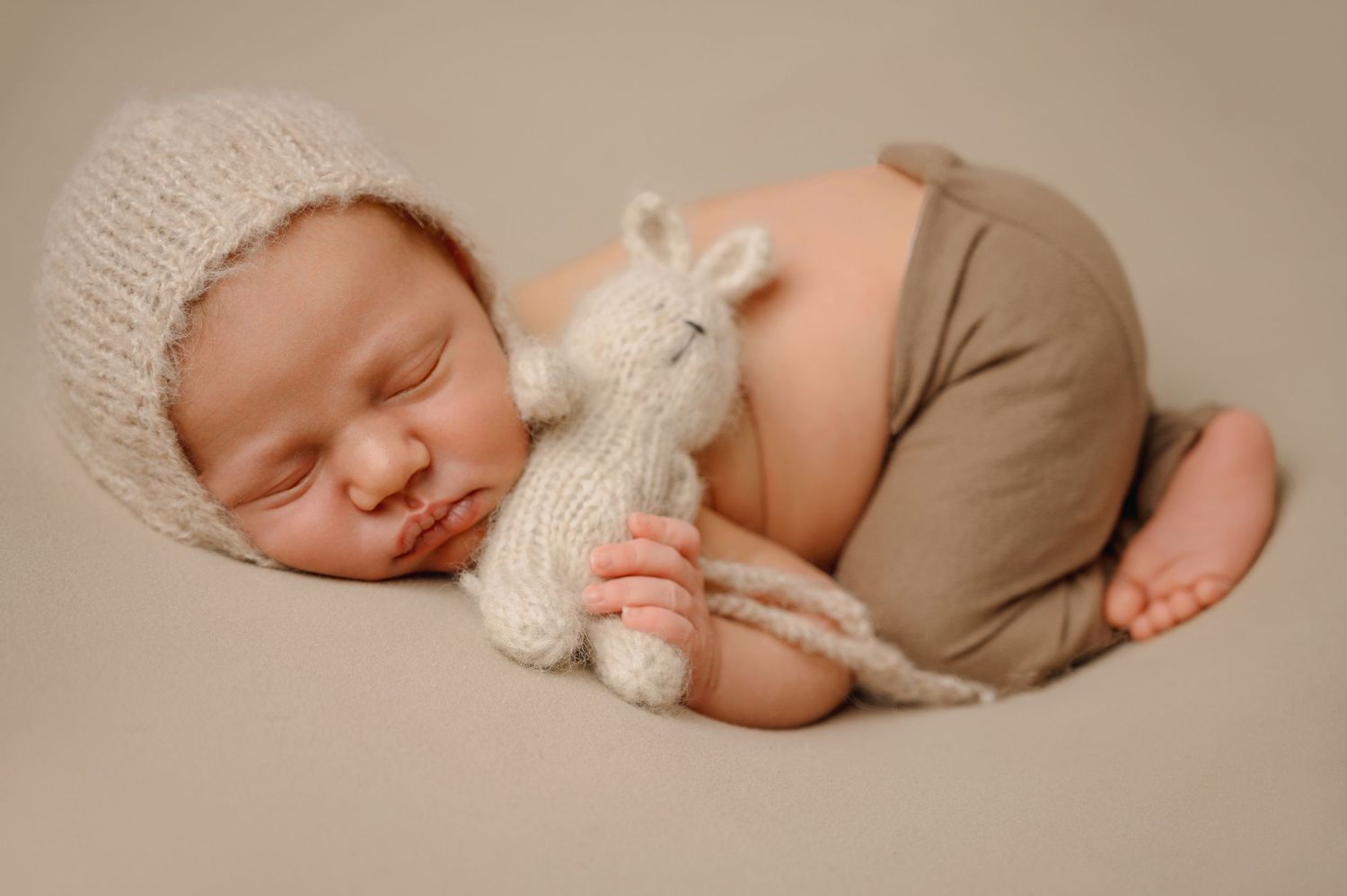A sleeping baby in a white knit hat cuddles with a stuffed bunny while wearing beige pants.