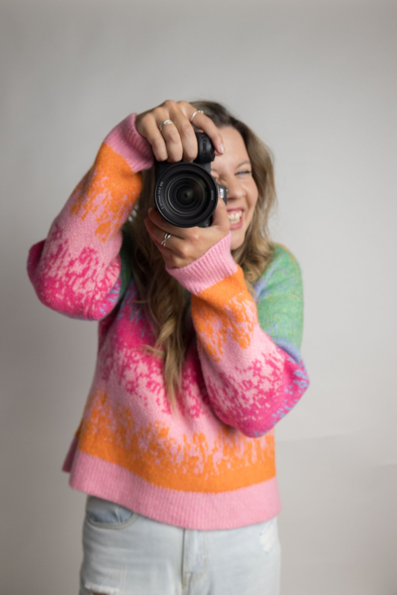 Photographer wearing colorful tie-dye style sweater in pink, orange and green holds up camera against light gray background.