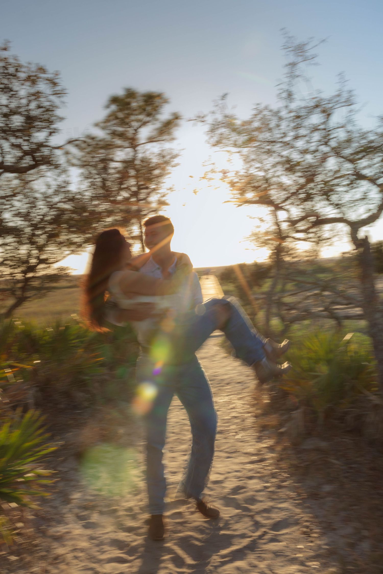 Couple spinning joyfully on sandy path surrounded by trees in warm golden sunset light with lens flare.