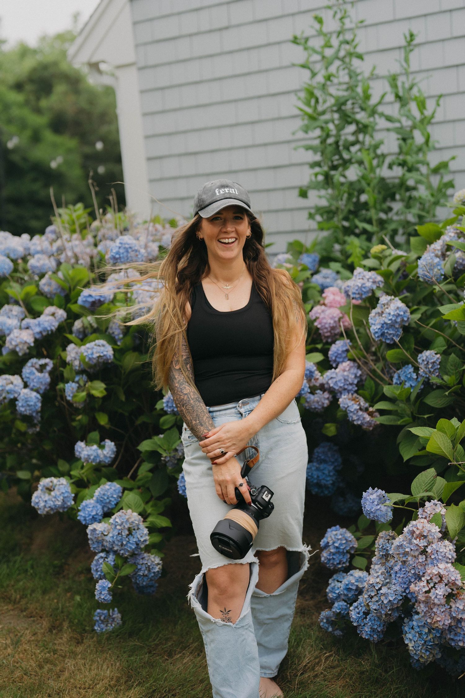 A smiling woman with long hair holds a camera while standing among blooming blue hydrangeas beside a gray building.