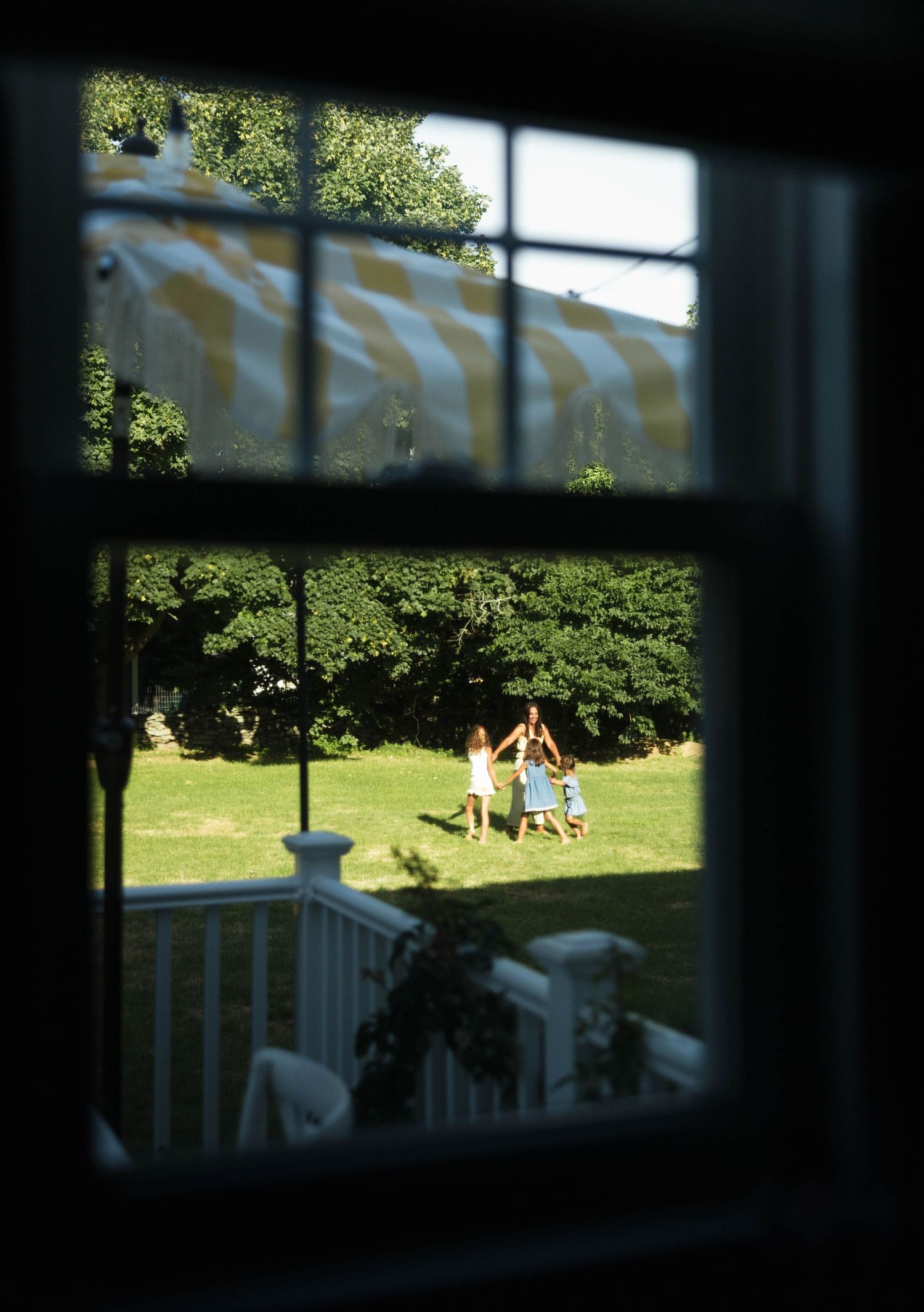 A family plays together in a sunny green backyard, viewed through a vintage window with white porch railing.