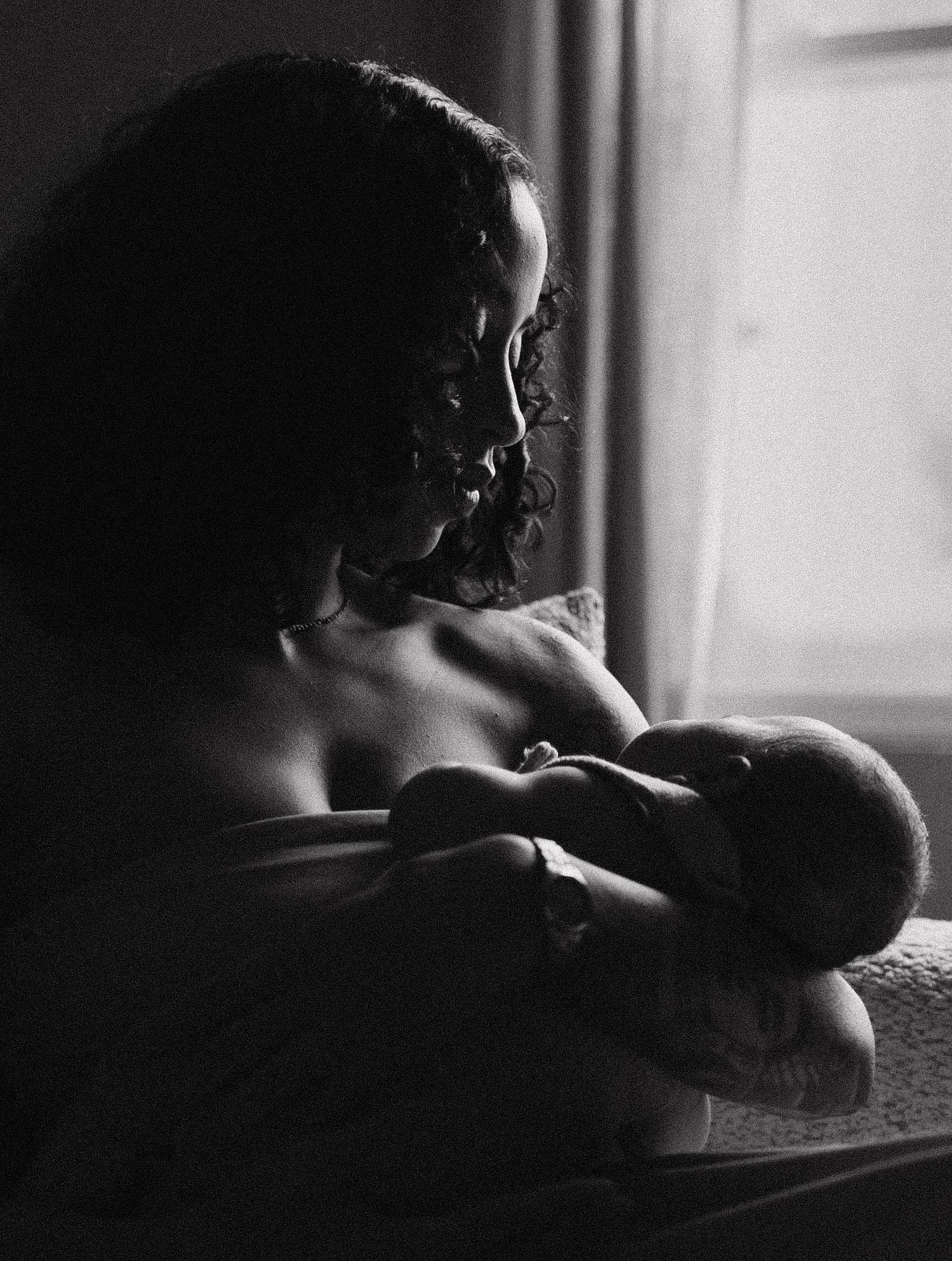 Black and white portrait of a mother breastfeeding newborn baby by a softly lit window.