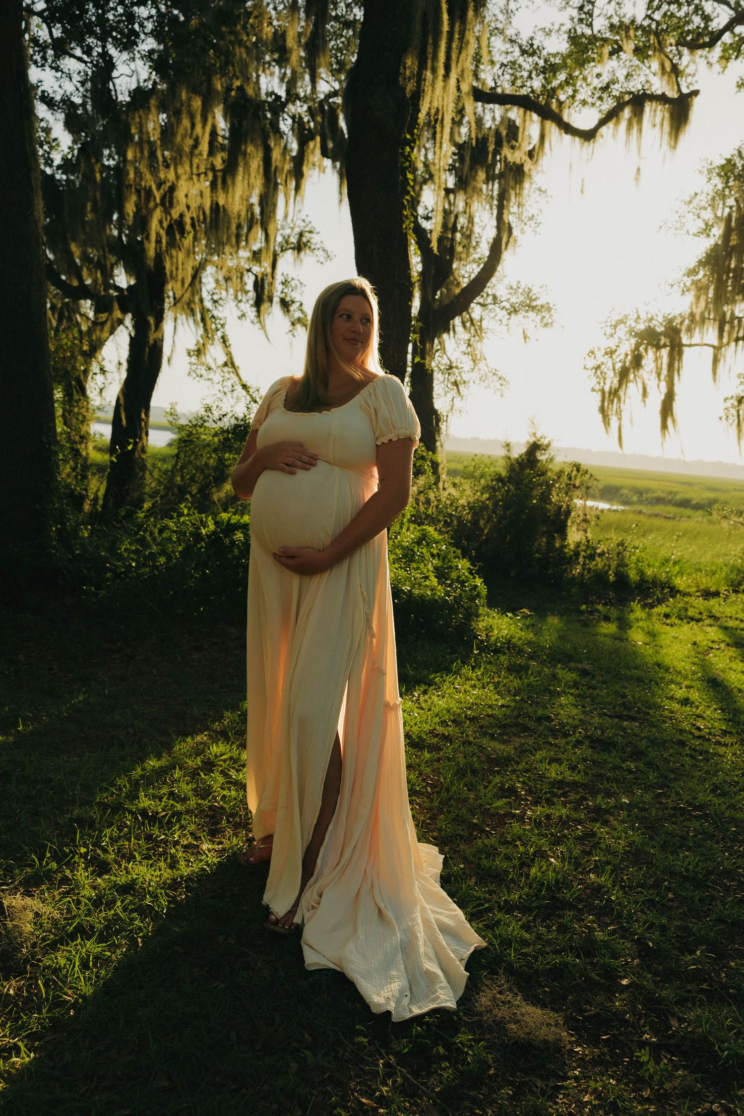 Pregnant woman in elegant cream maternity gown stands under Spanish moss trees at golden hour.