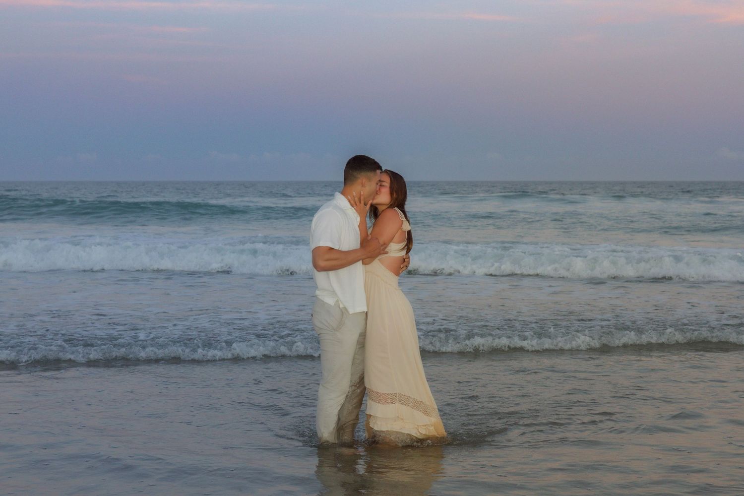 Couple sharing romantic kiss standing in ocean waves at sunset, woman in white wedding gown.
