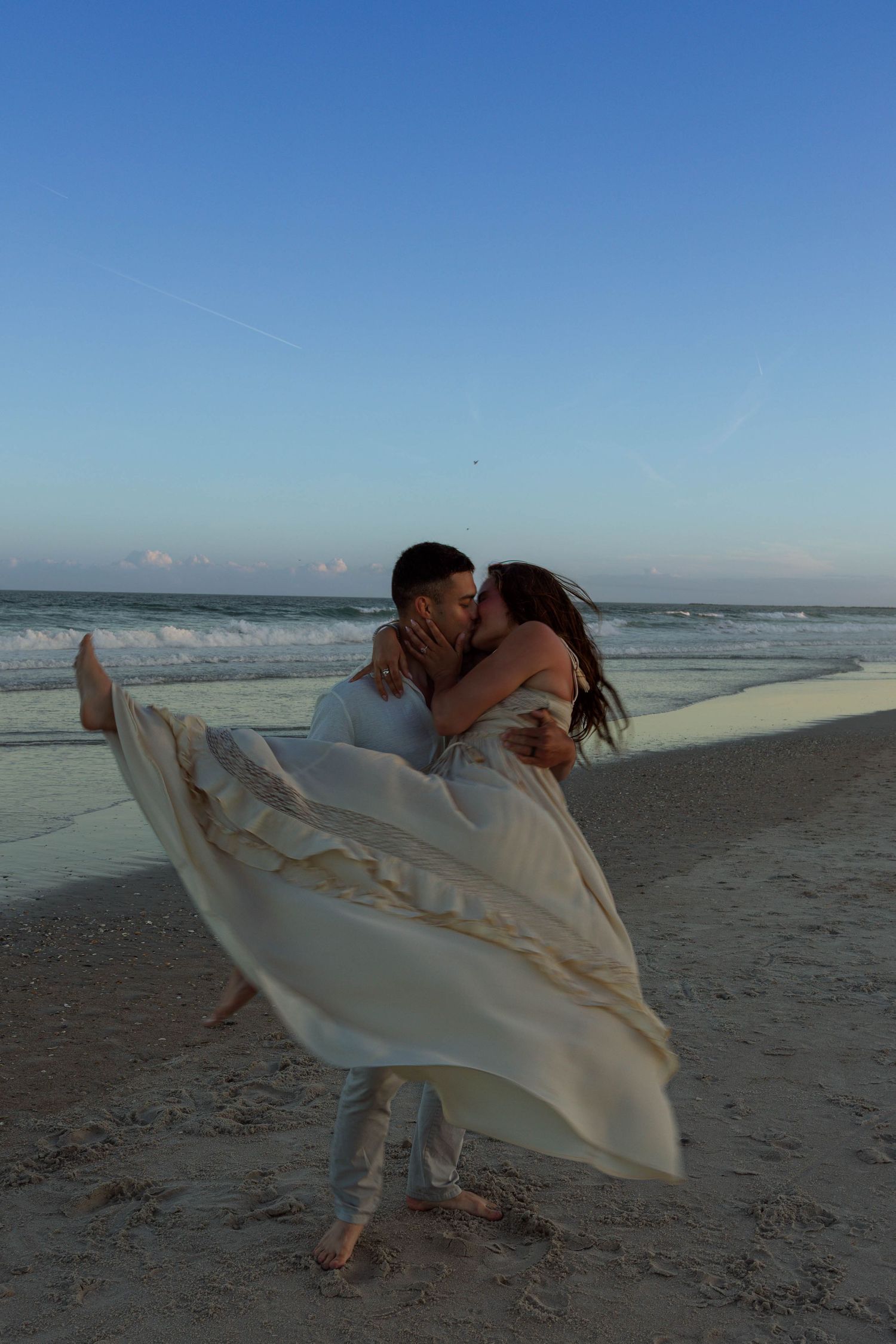 Groom lifts bride in flowing white dress for romantic kiss on sandy beach at dusk by ocean waves.