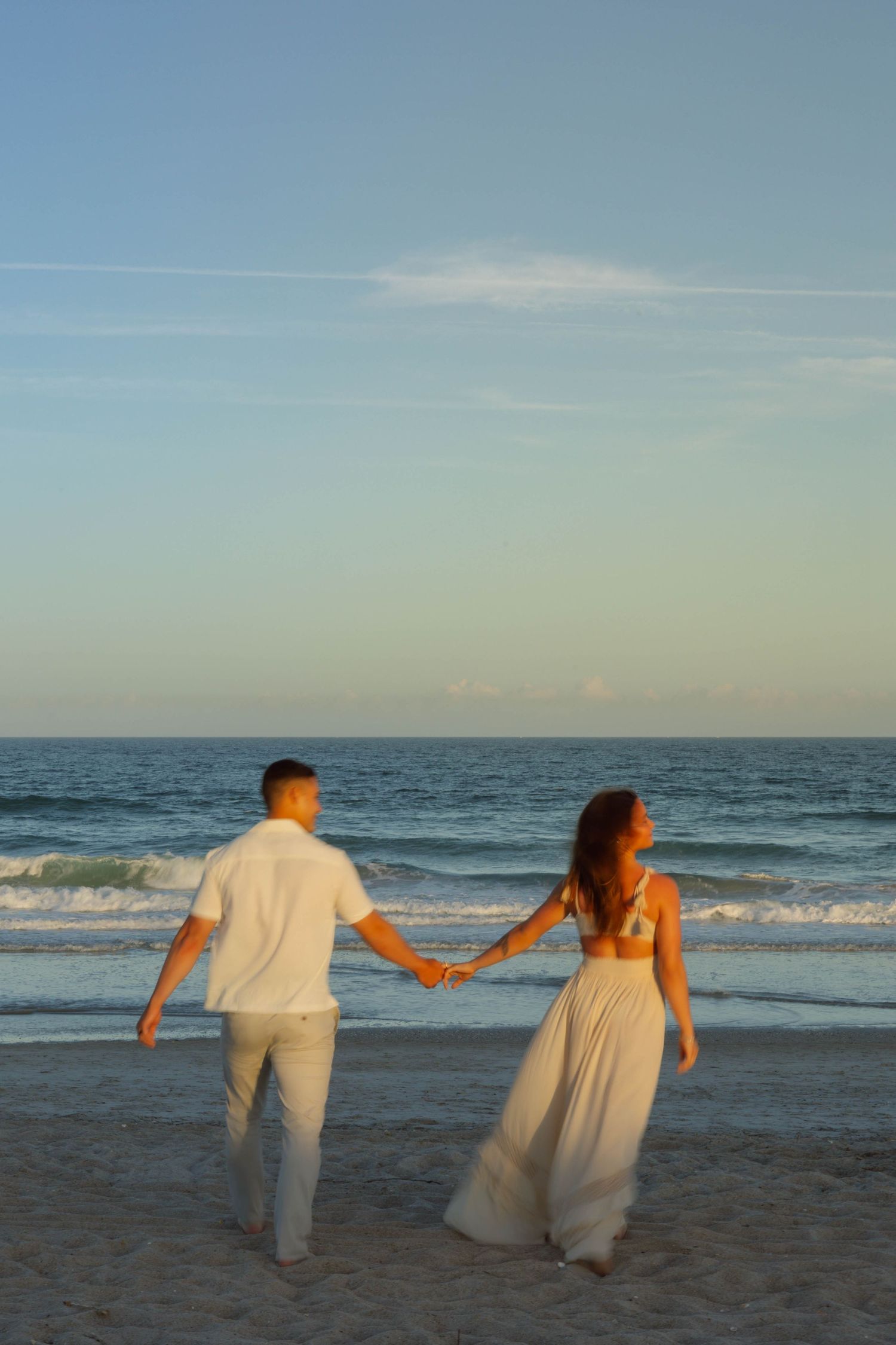 Couple in white wedding attire holds hands walking toward ocean waves at golden hour beach sunset.